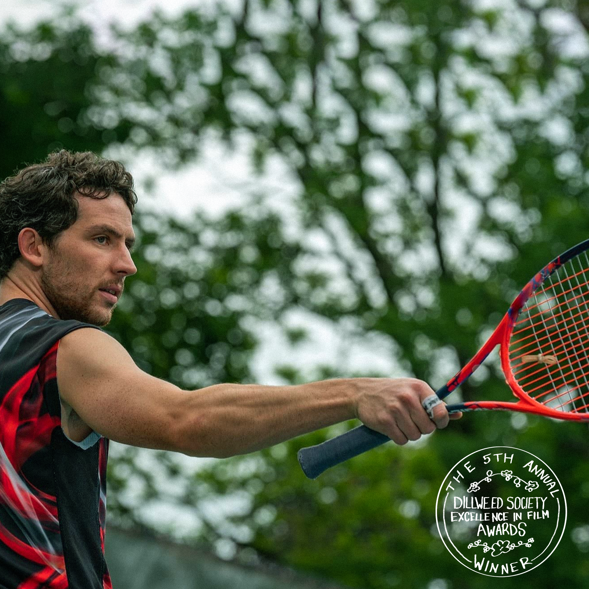 A man playing tennis outdoors with trees in the background, holding a red tennis racket, wearing a sleeveless athletic shirt, and a circular award badge in the bottom right corner indicating he is a winner of the 5th Annual Dillweed Society Excellence in Film Awards.
