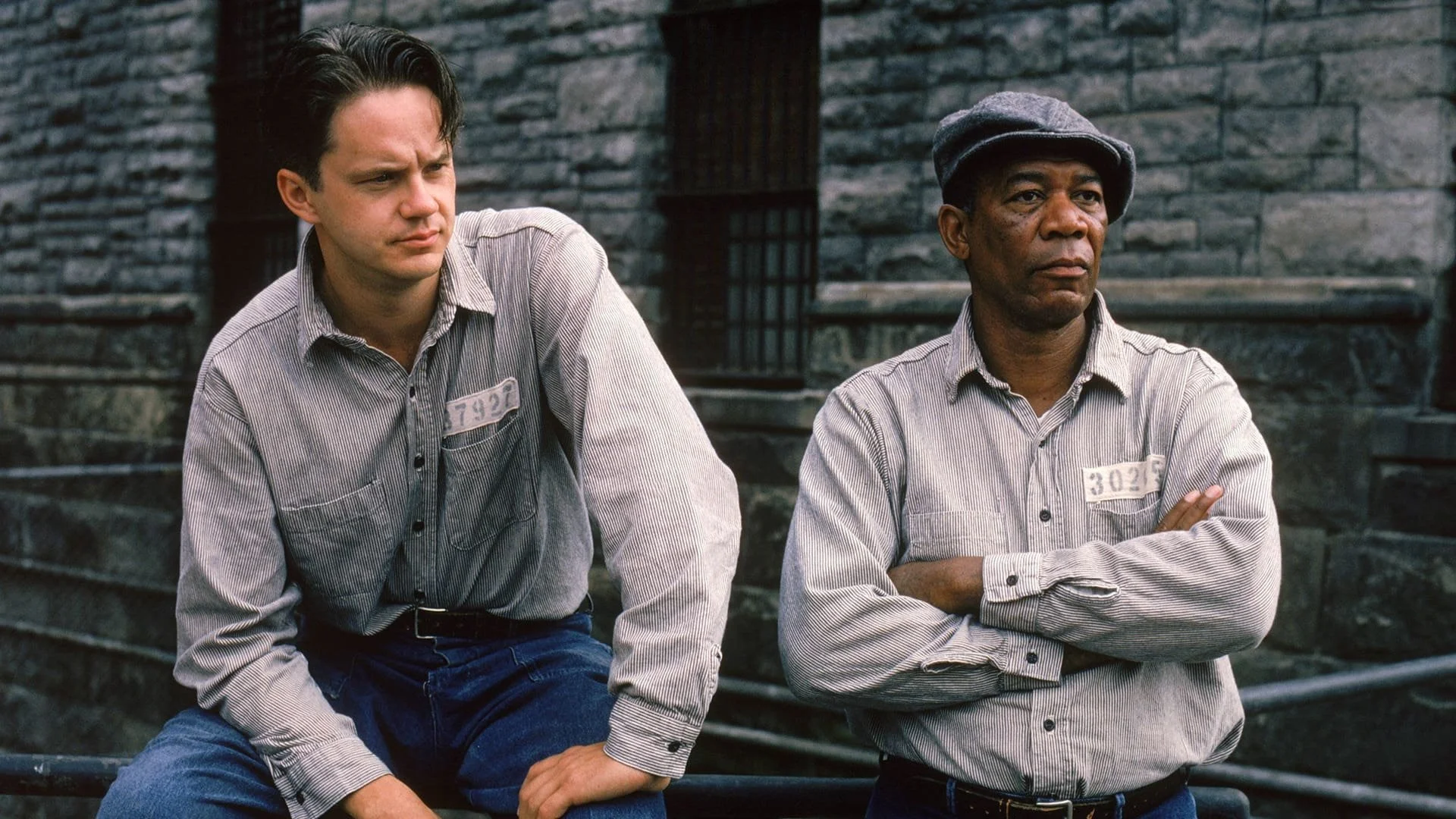 Two men, one younger and one older, standing against a stone building. The younger man has dark hair, a striped shirt, and jeans, sitting with an unsure expression. The older man has a gray cap, striped shirt, and crosses his arms, looking serious.