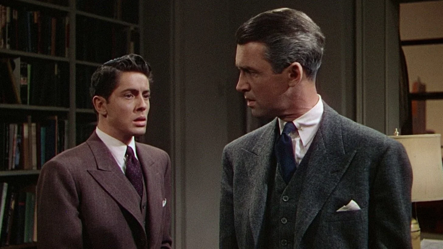 Two men in suits having a serious conversation indoors, with bookshelves and a lamp in the background.