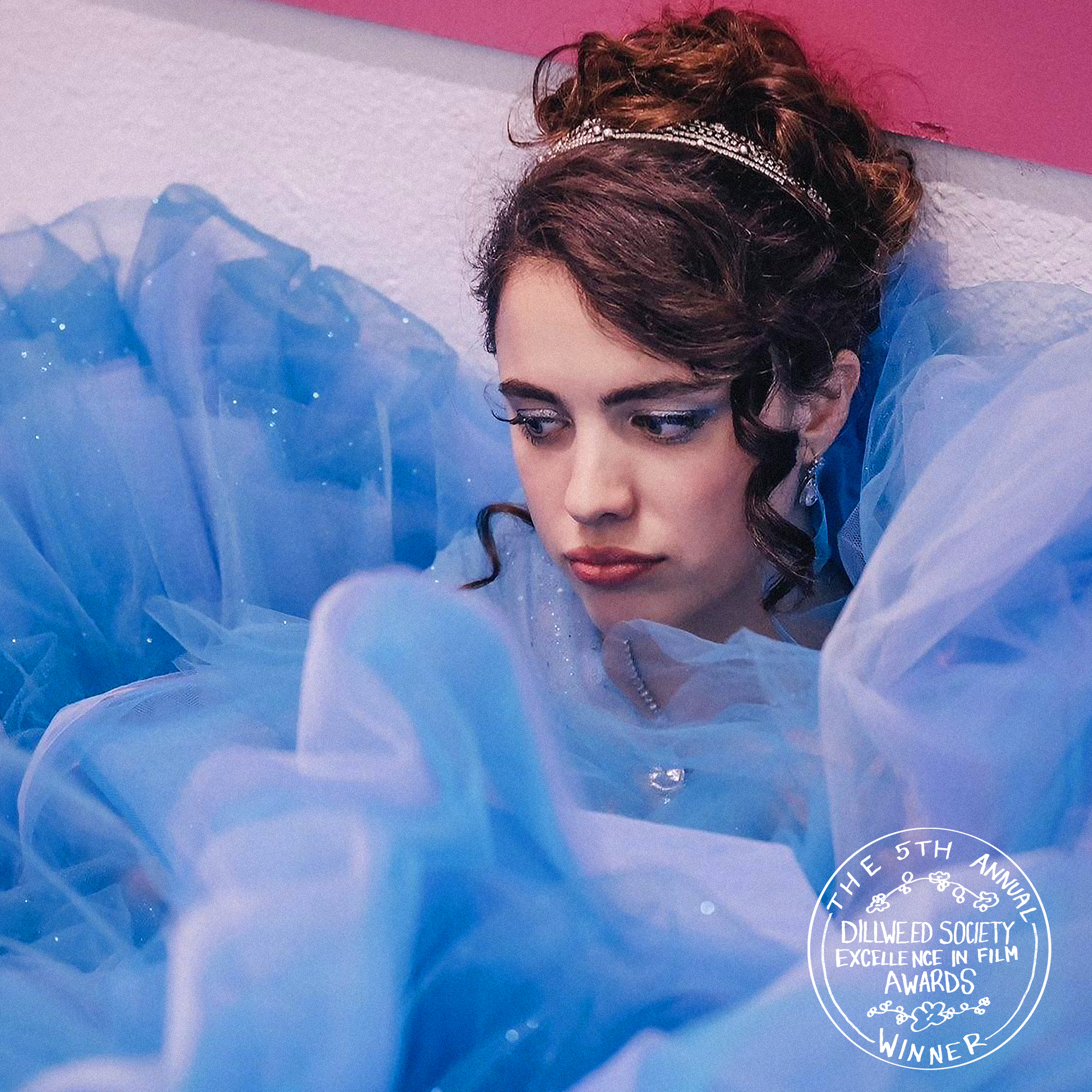 Young woman dressed as a princess, wearing a tiara and blue gown, looking down thoughtfully. A badge in the bottom right indicates she is a winner of the 5th Annual Dillweed Society Excellence in Film Awards.