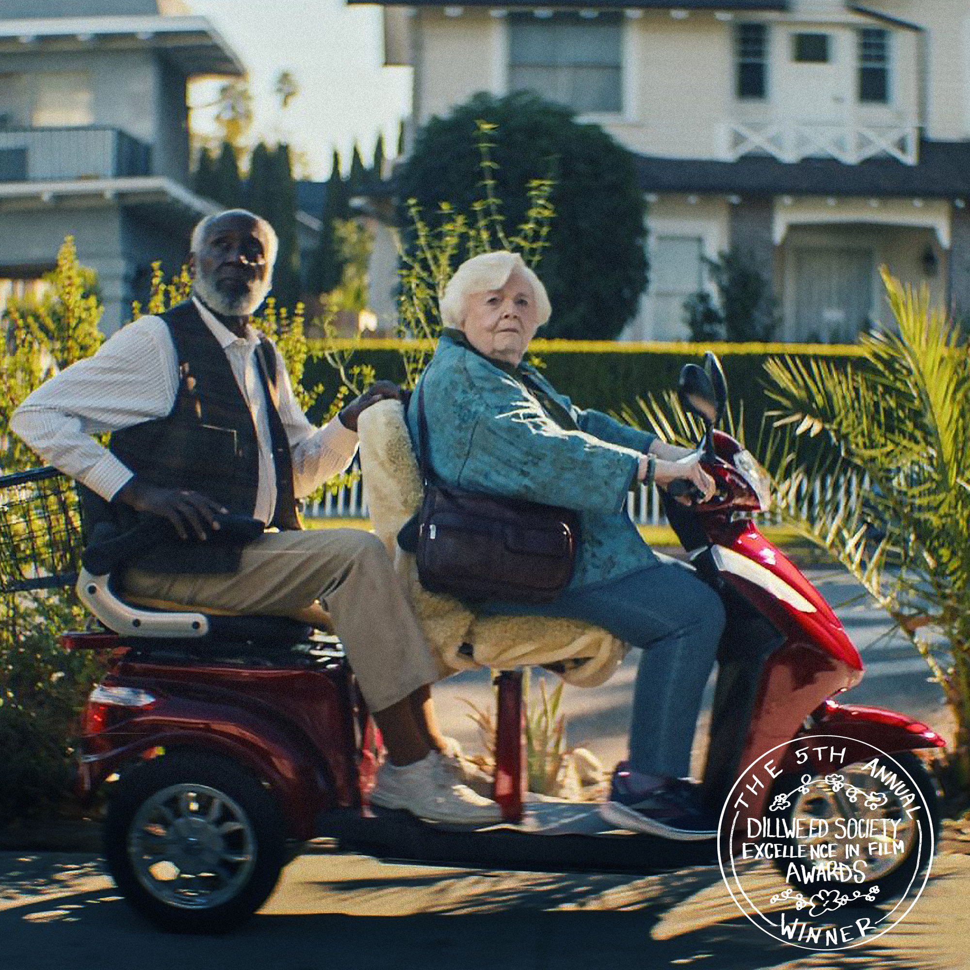 An elderly woman rides a red mobility scooter with a man sitting behind her. The woman has white hair and wears a teal jacket. The man has gray hair and a beard, and wears a white shirt with a black vest. The background shows a residential neighborhood with houses and greenery. There is a white circular emblem in the bottom right corner that reads 'The 5th Annual Dillweed Society Excellence in Film Awards Winner.'