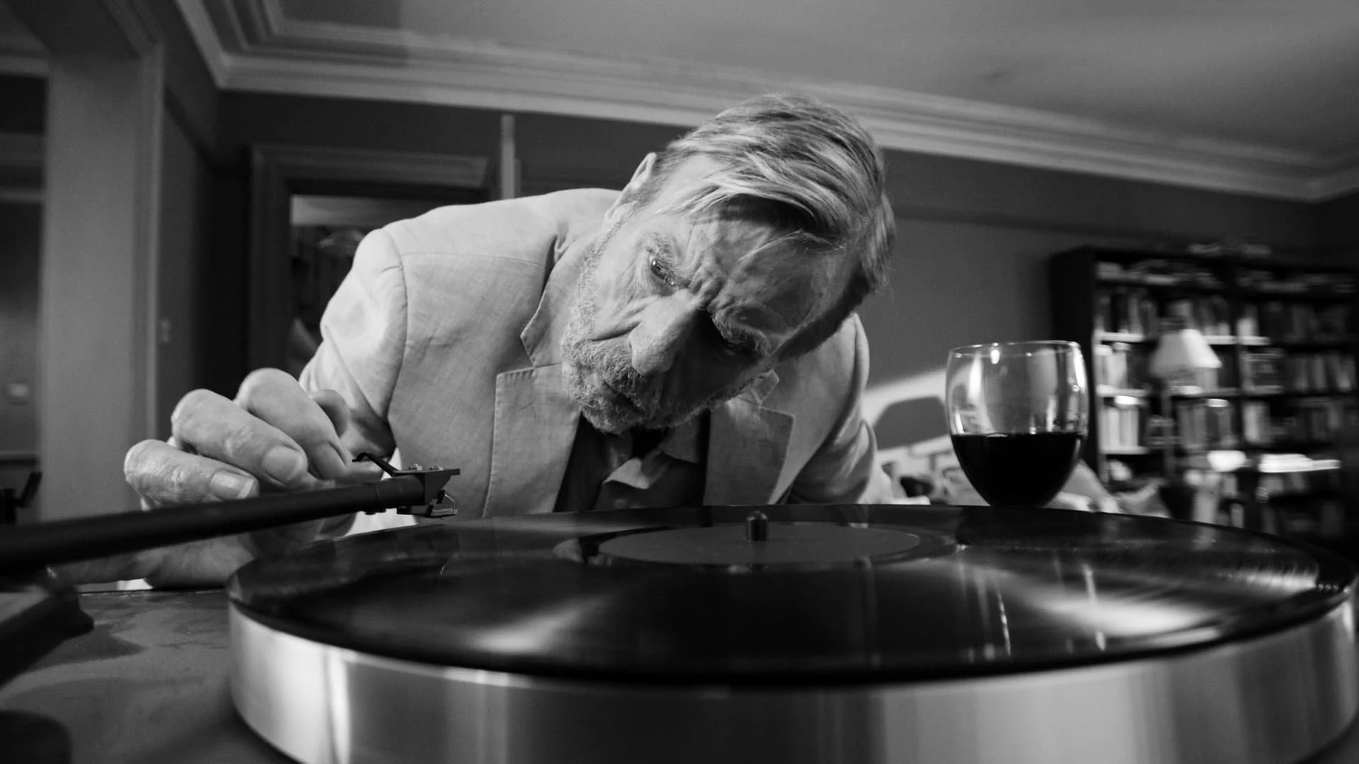 An elderly man with gray hair and beard, wearing a light-colored blazer, leaning over a record player, with a glass of red wine and a bookshelf in the background.
