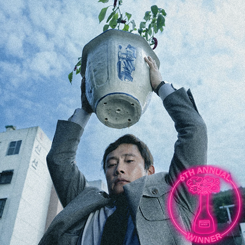 Close-up of a woman's face in profile, smiling with visible teeth, showing brown eyes, smooth skin, and dark hair, with a dark background and an award emblem in the bottom right corner.