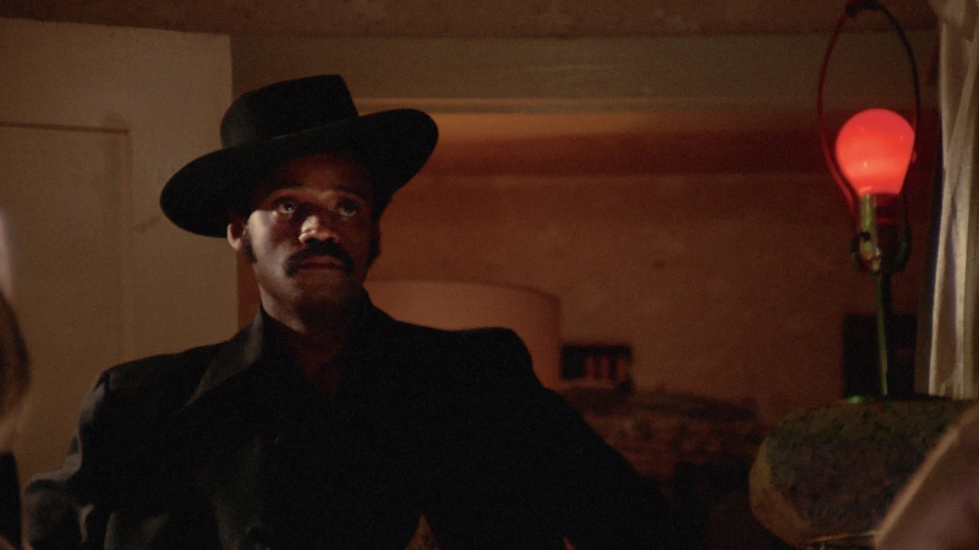 A man dressed in a black suit and wide-brimmed black hat sitting at a table in a dimly lit room with a red lamp on the side.