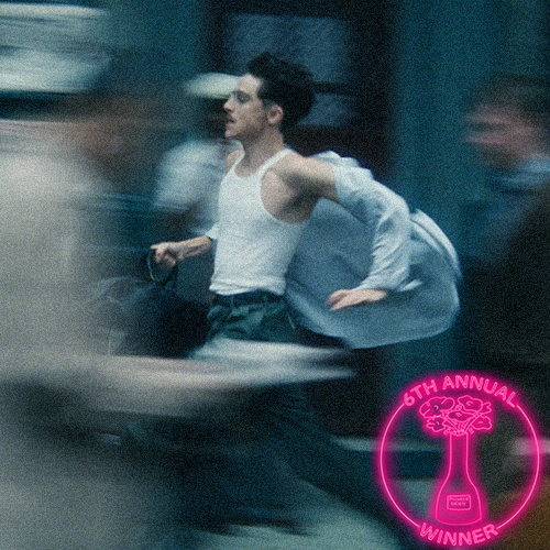 Two men having a conversation at night with city lights in the background. One has curly hair and glasses; the other has a bandana around his head and a beard. There is a logo in the bottom right corner indicating an award for excellence in film.