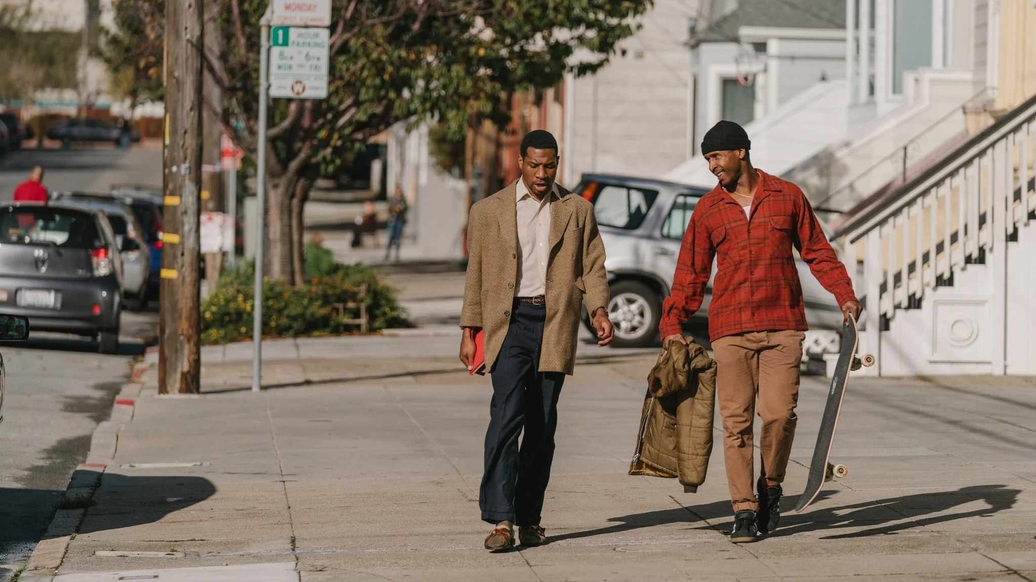 Two men walk along a city sidewalk, one dressed in a brown coat and the other in a red plaid shirt, carrying a skateboard and a bag.