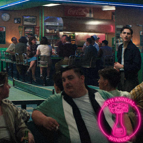 A group of people dressed in colorful costumes, including cowboy hats and traditional attire, gathered in a dark room with a spotlight overhead. They appear to be participating in a presentation or ceremony, with one individual speaking into a microphone. There is a circular badge in the lower right corner that reads 'The 5th Annual Dillweed Society Excellence in Film Awards Winner.'