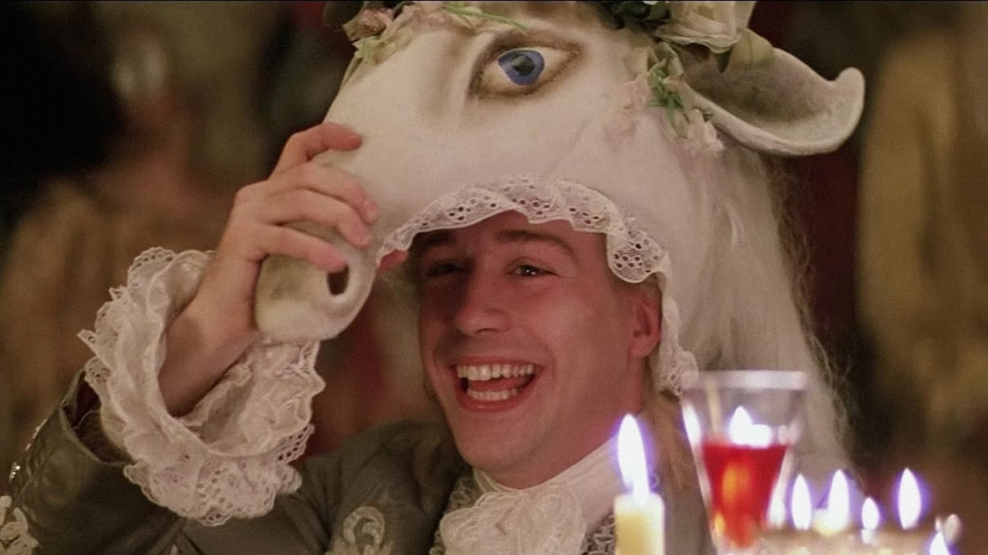 A man smiling and laughing while holding a mask of a horse's face on his head, wearing a lace-trimmed wig and Victorian-style clothing, with candles in glasses in front of him.