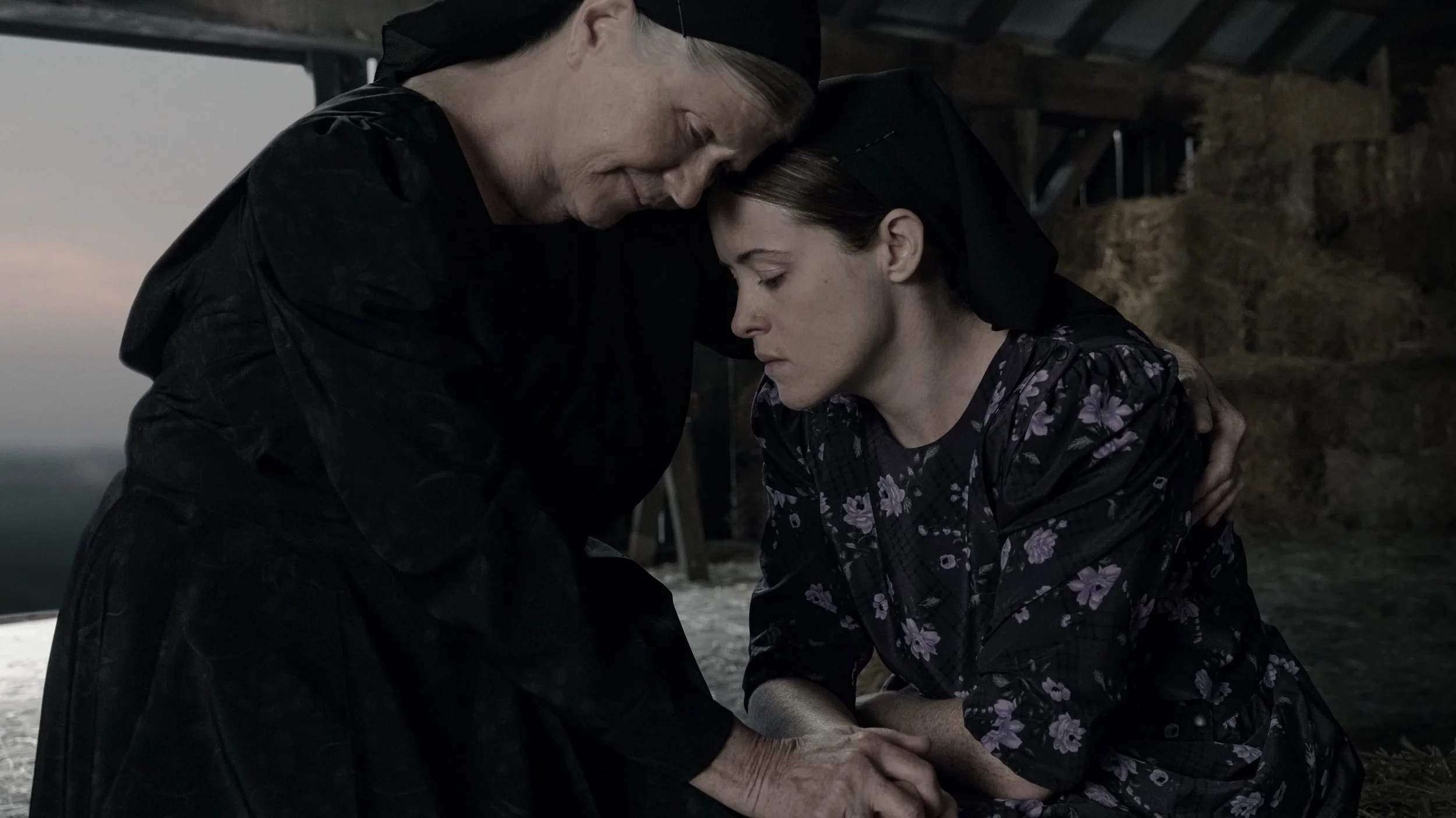 Two women with veiled hair lean their heads together with closed eyes, holding hands in a prayer or comforting gesture inside a rustic barn or shed.