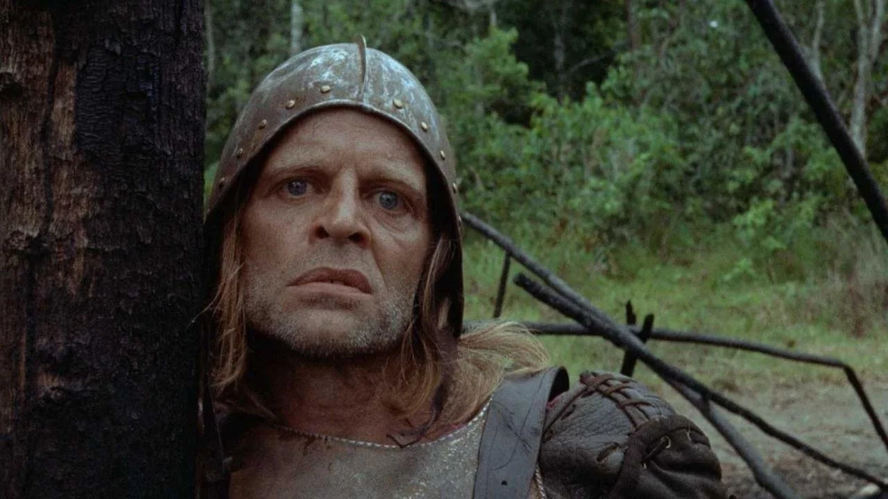 A man dressed as a medieval knight with a helmet, looking anxiously behind a tree in a forest.