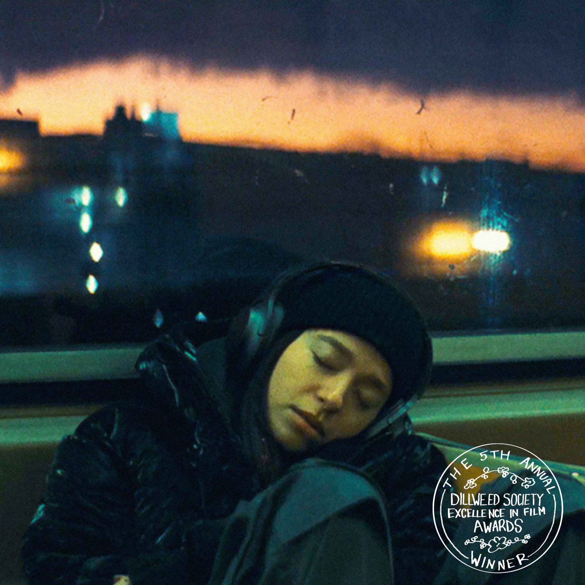 A woman with closed eyes, wearing a black beanie and headphones, sleeping on a bus or train at night with city lights and a sunset visible through the window. A circular award badge in the bottom right corner indicates she is a winner of the 5th Annual Dillweed Society Excellence in Film Awards.
