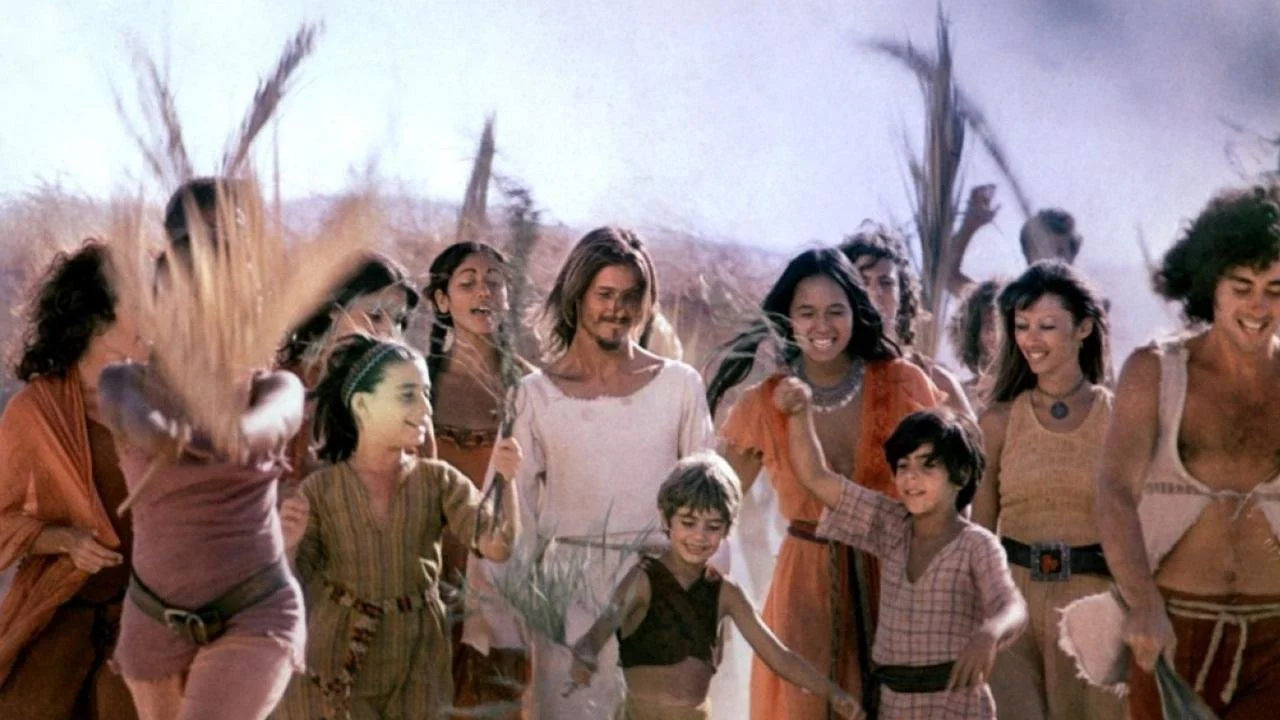 A group of smiling people, including children and adults, are outdoors in a desert landscape with dried plants and trees, possibly celebrating or enjoying a communal event.