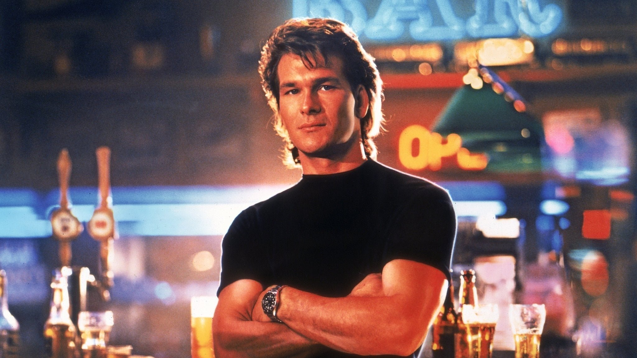A man with brown hair wearing a black t-shirt, arms crossed, standing in a bar with beer glasses and neon signs in the background.