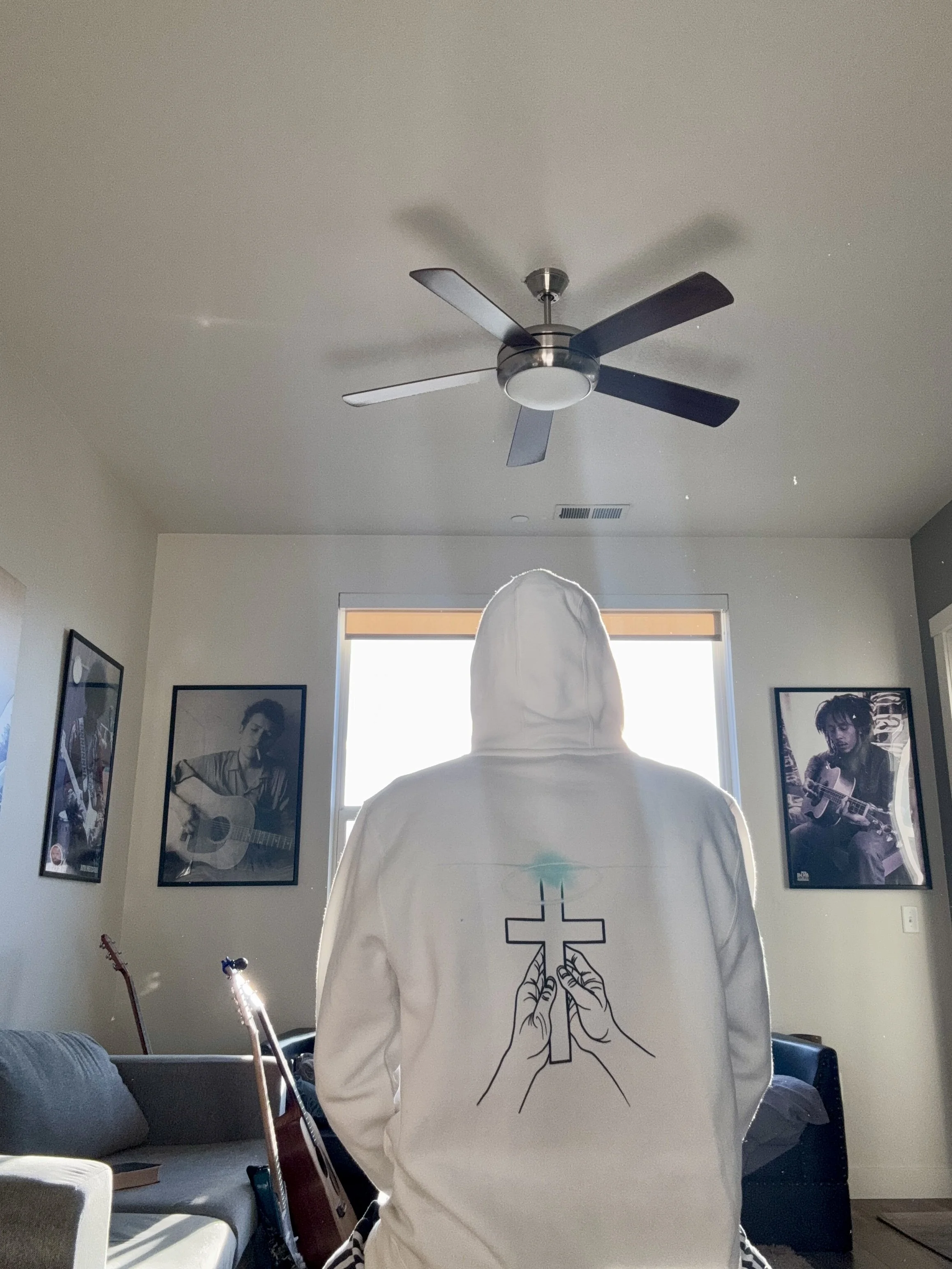 Person wearing a white hoodie with a cross and hands holding a candle printed on the back, sitting in a living room near a window. The room has framed photos of musicians on the walls, an acoustic guitar, and a ceiling fan.