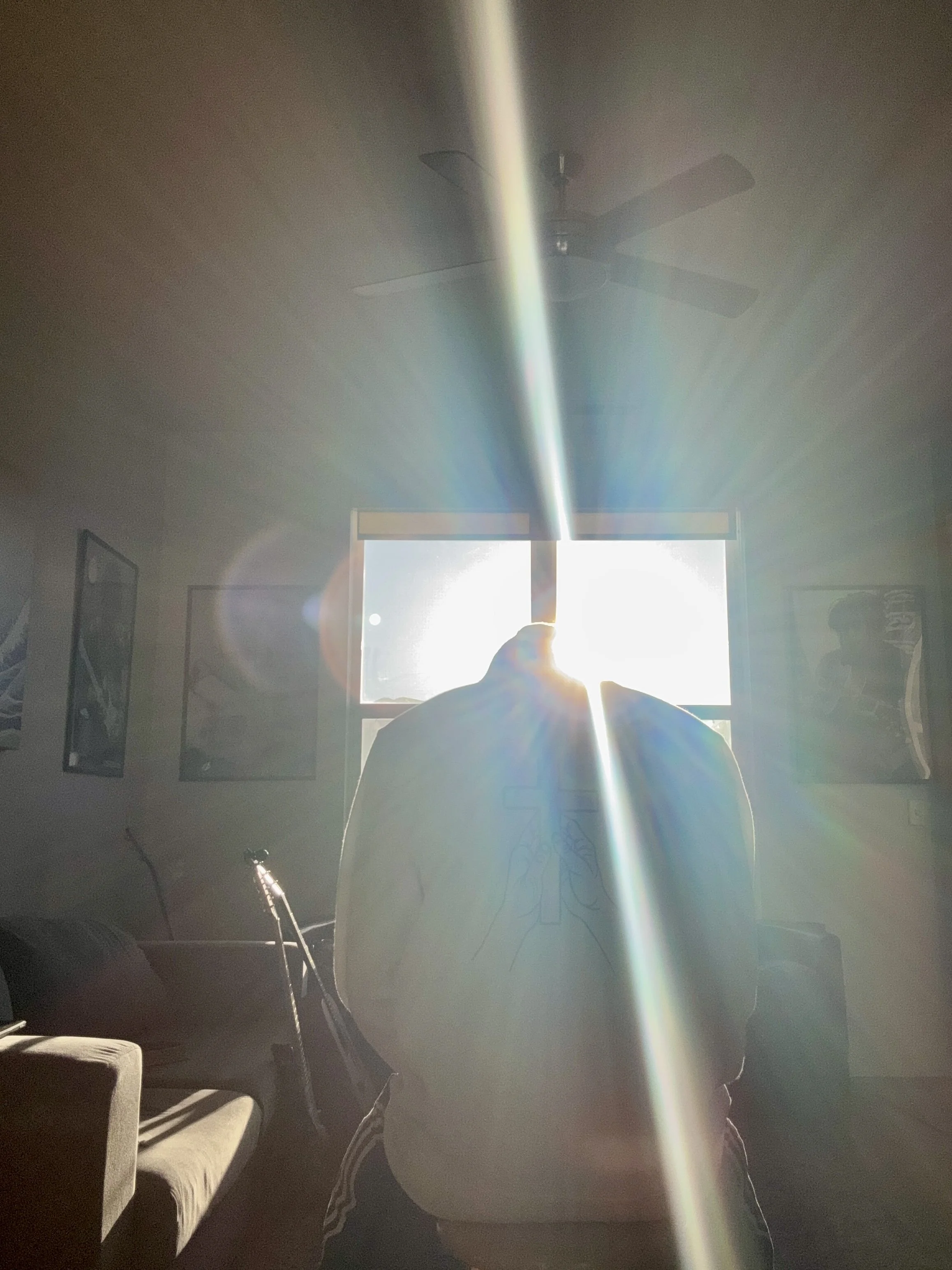 Person sitting in a room with sunlight streaming through a window, creating a lens flare and backlighting the person's silhouette.