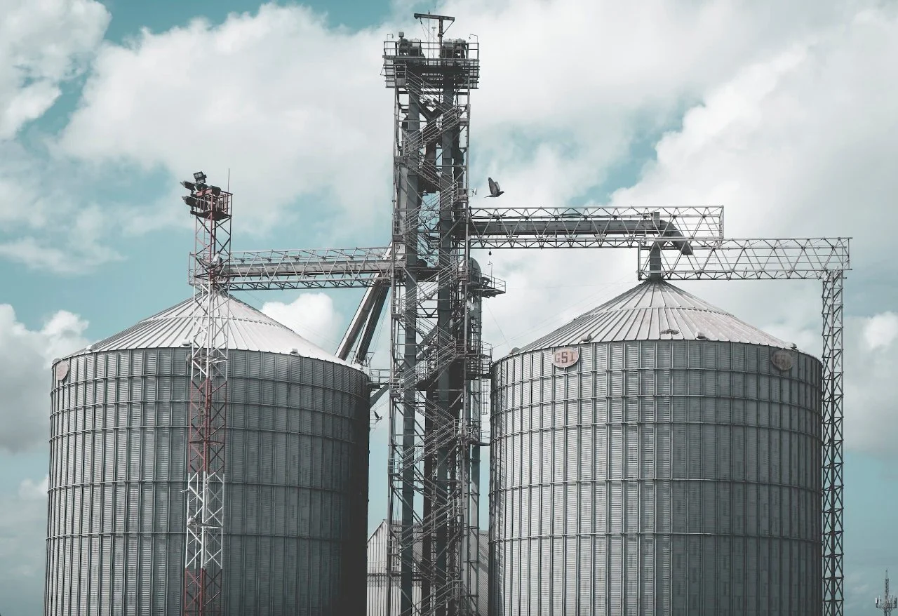 Two large metal grain silos with a metal structure in the middle connecting them, under a cloudy sky. A bird is flying near the structure.