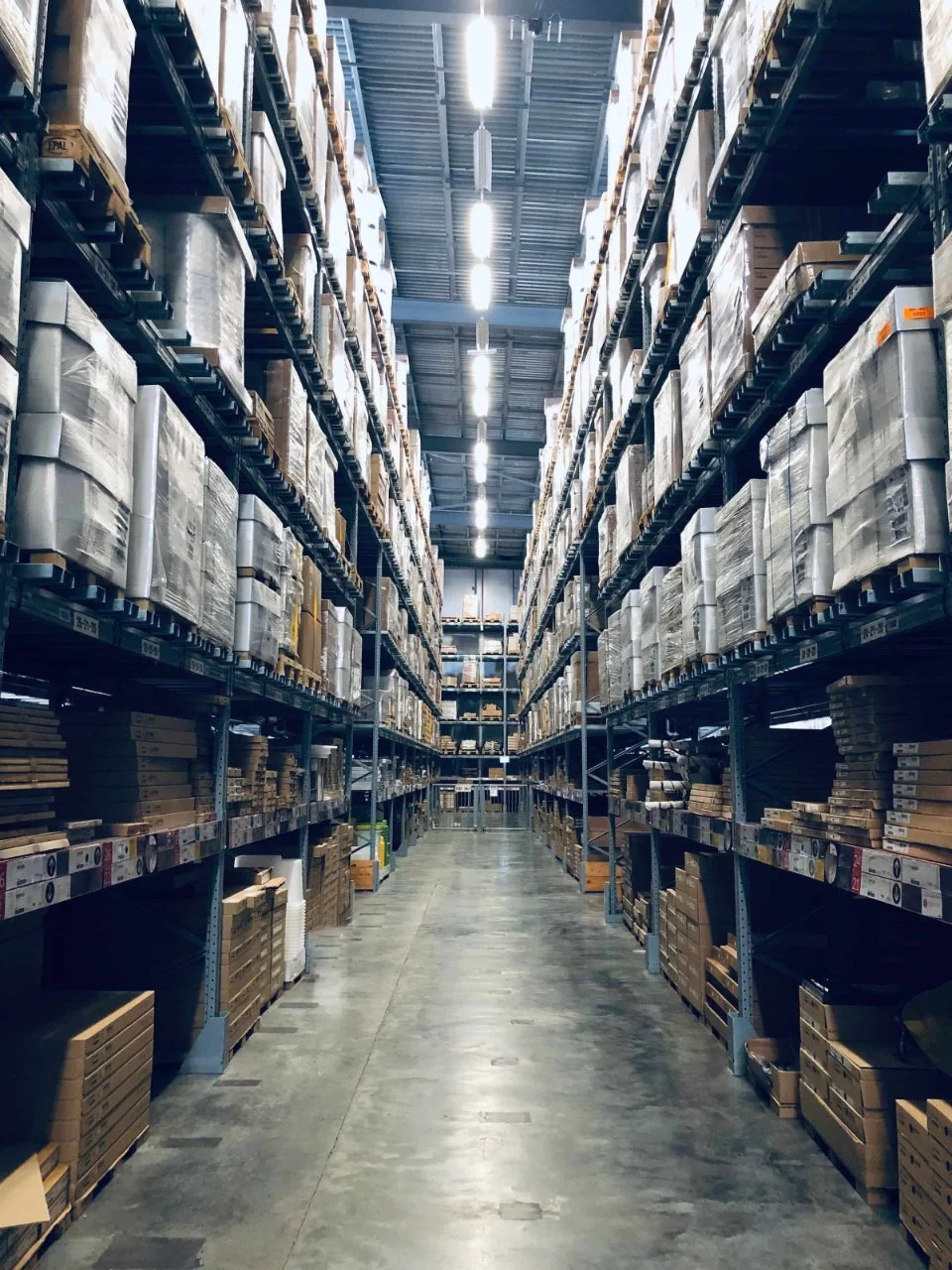 Warehouse aisle with tall shelves filled with boxes and packaging materials, illuminated by overhead lighting.