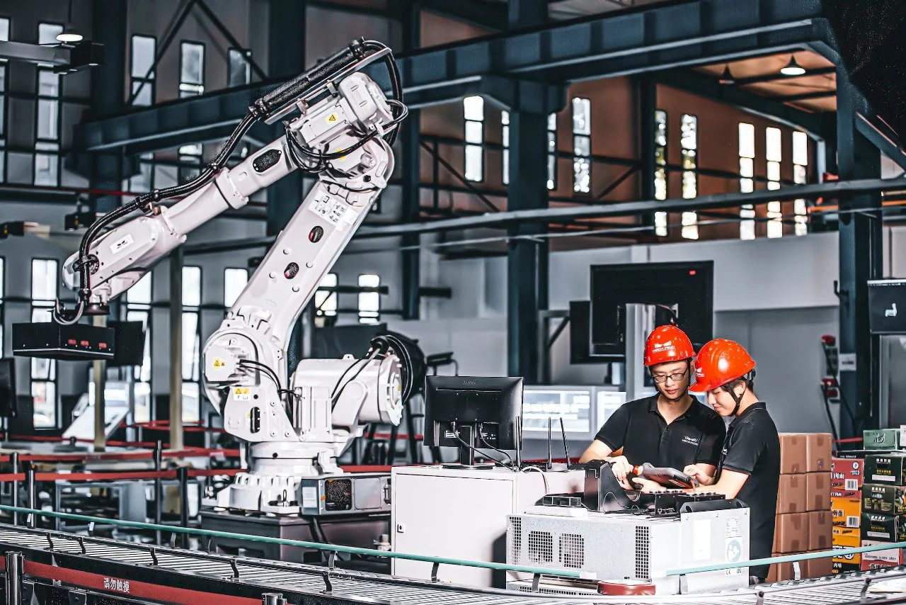 Two workers wearing red safety helmets and black shirts operate a robotic arm in an industrial setting, with machinery and boxes around them.