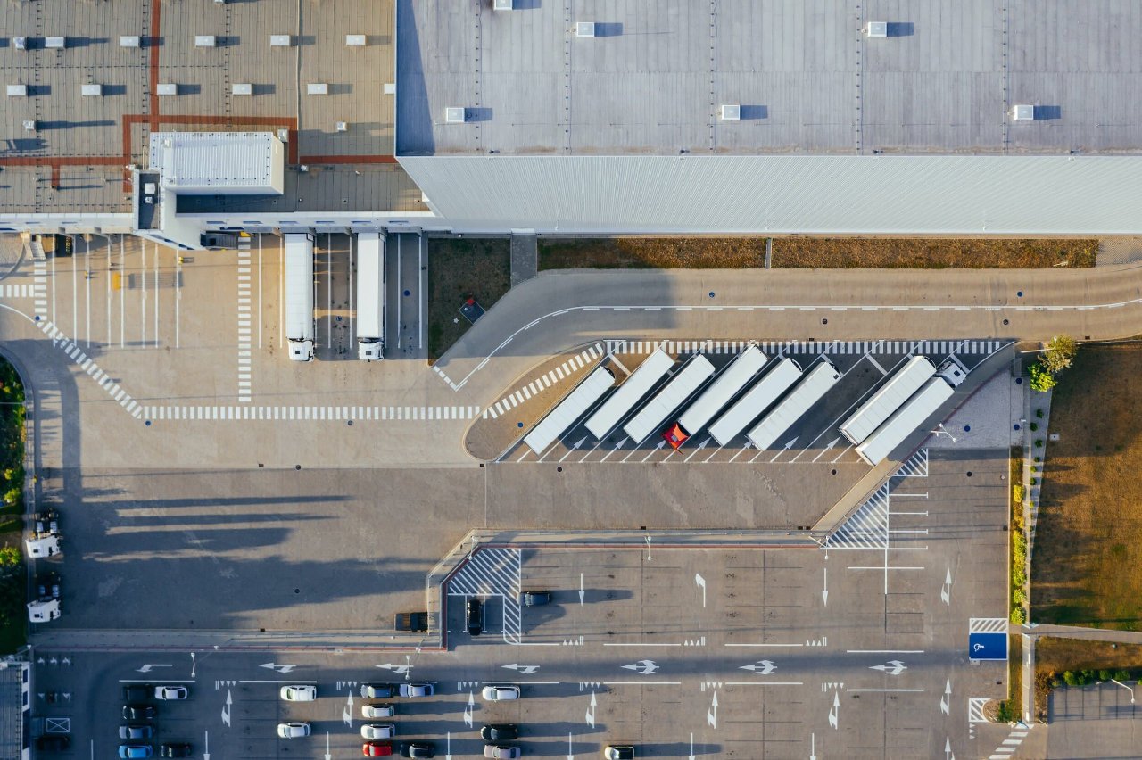 Aerial view of a large parking lot with cars, trucks, and a loading dock area next to a building, with roadways and marked parking spaces.