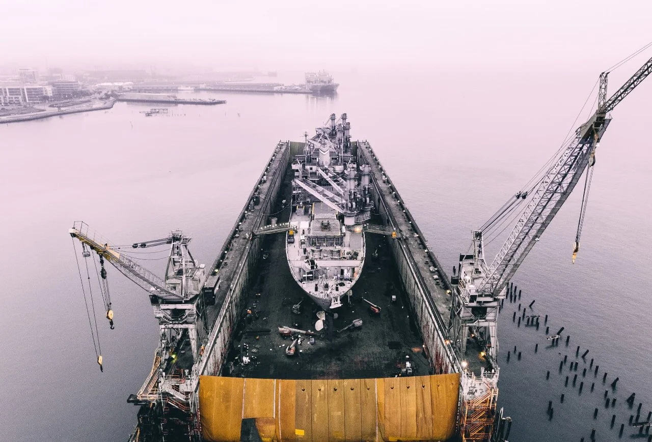 A large ship is in a shipyard with two cranes on either side, preparing for maintenance or construction, with water and some buildings visible in the background.