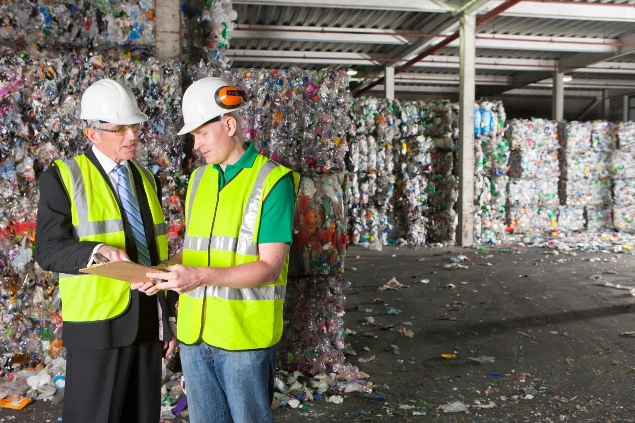 Two workers wearing safety helmets and high-visibility vests discussing in a recycling facility with large stacks of compressed plastic bottles in the background.