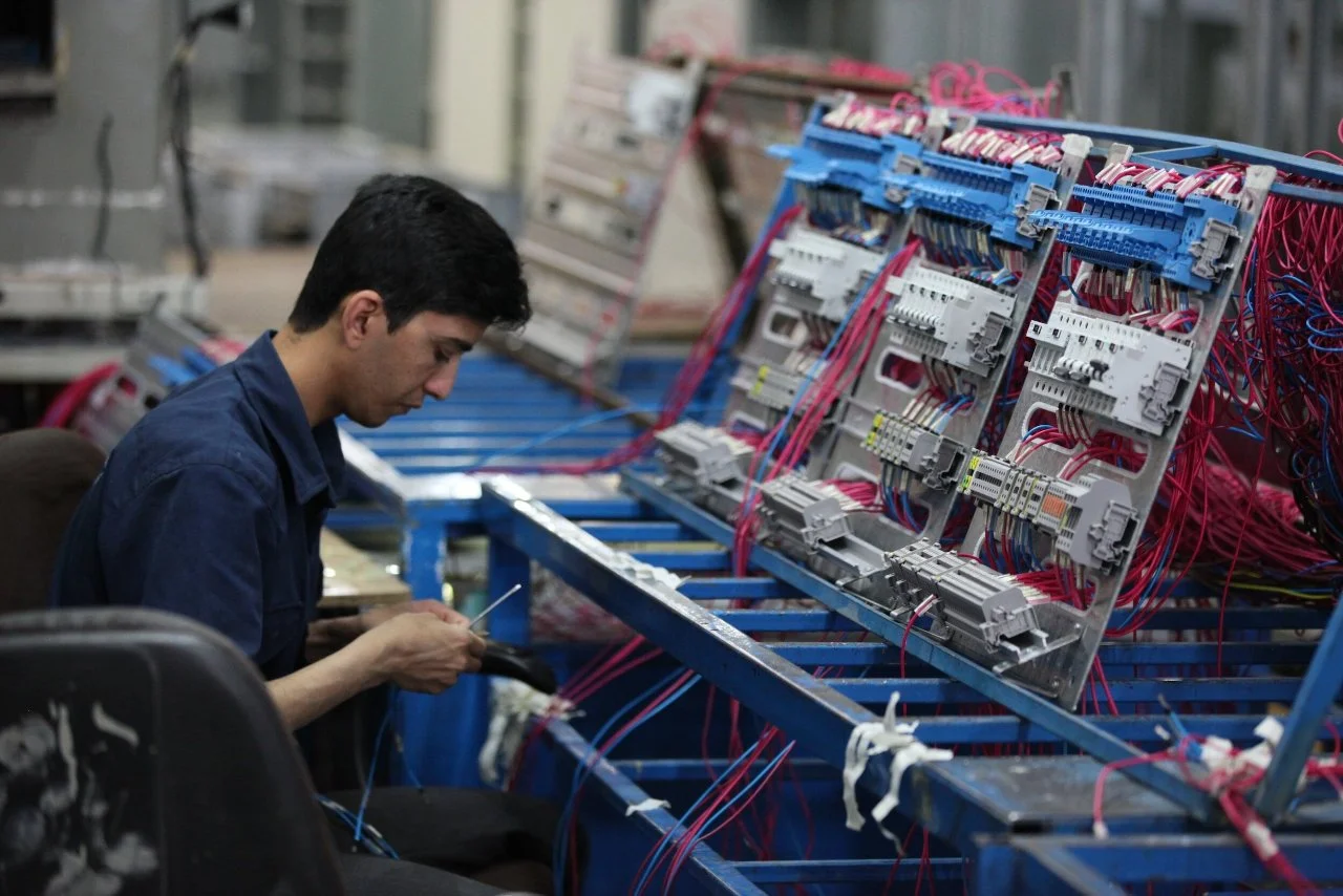 A man working on electrical wiring with circuit panels and pink wires on a blue workbench in an industrial setting.