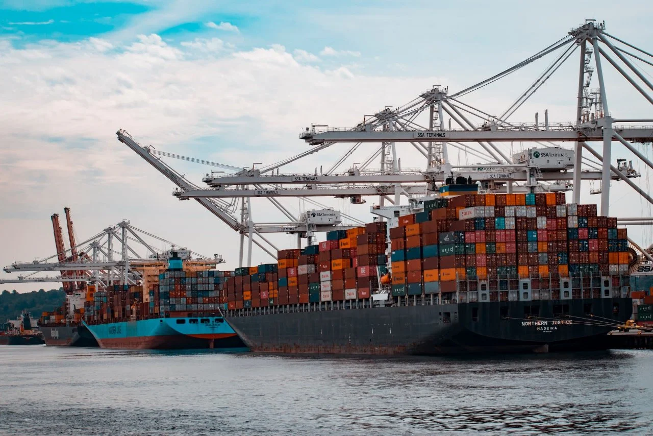 Cargo ships docked at a port with large cranes loading containers, water in foreground, cloudy sky above.