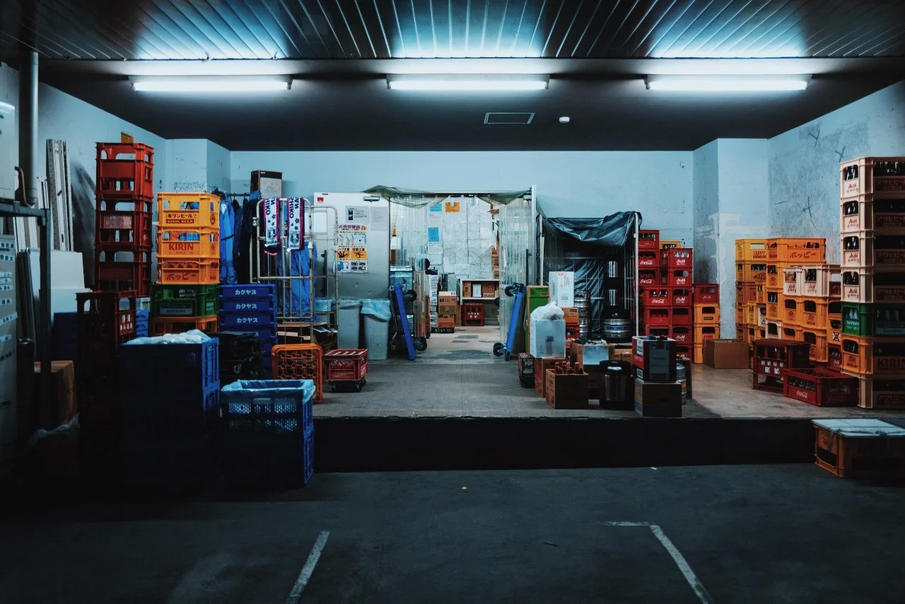 Underground storage area with stacks of orange, red, green, and blue plastic crates, some stacked on shelves, others on the floor, and a small open kitchen or storage space with equipment and supplies visible in the background.