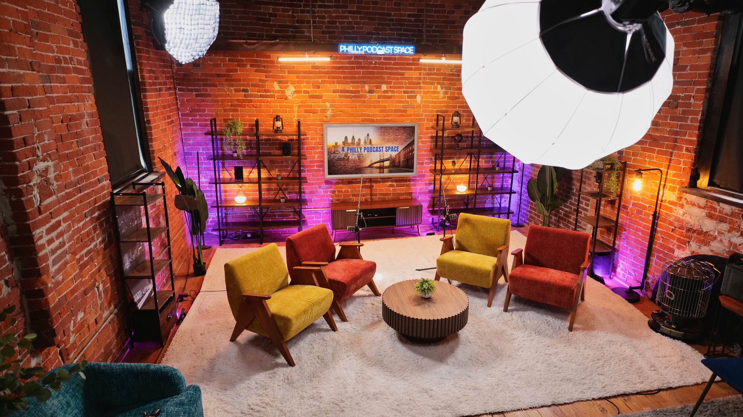 A cozy podcast recording studio with a brick wall background, colorful armchairs arranged around a round coffee table on a white rug, large studio lights hanging above, shelves with decorative items, and a flat screen TV displaying 'PHILLY PODCAST SP