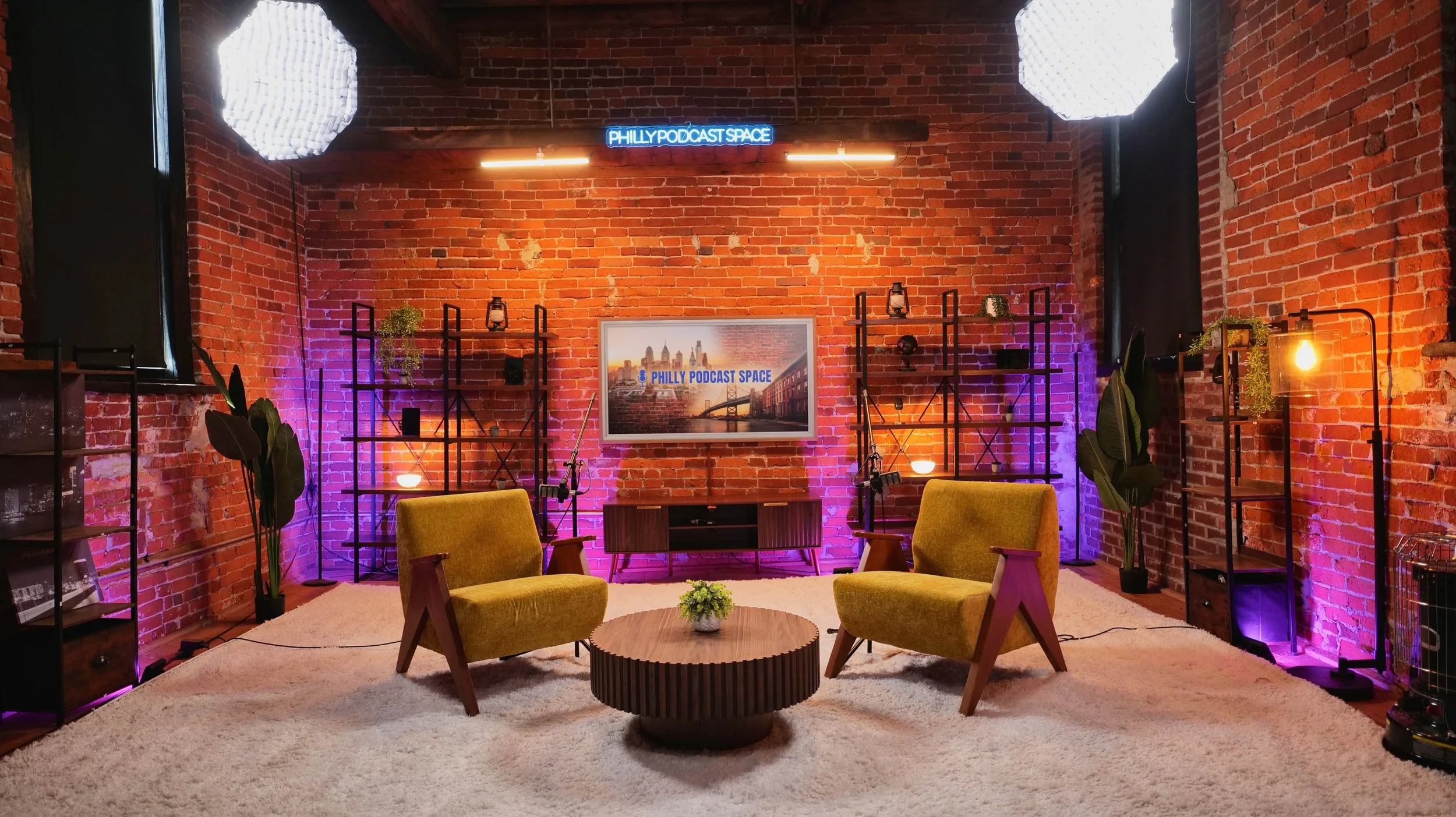 Interior of a podcast recording studio with brick walls, two yellow armchairs, a round wooden coffee table with a small plant, shelves with plants and decor, purple and orange lighting, and a large screen that says 'Philly Podcast Space'.