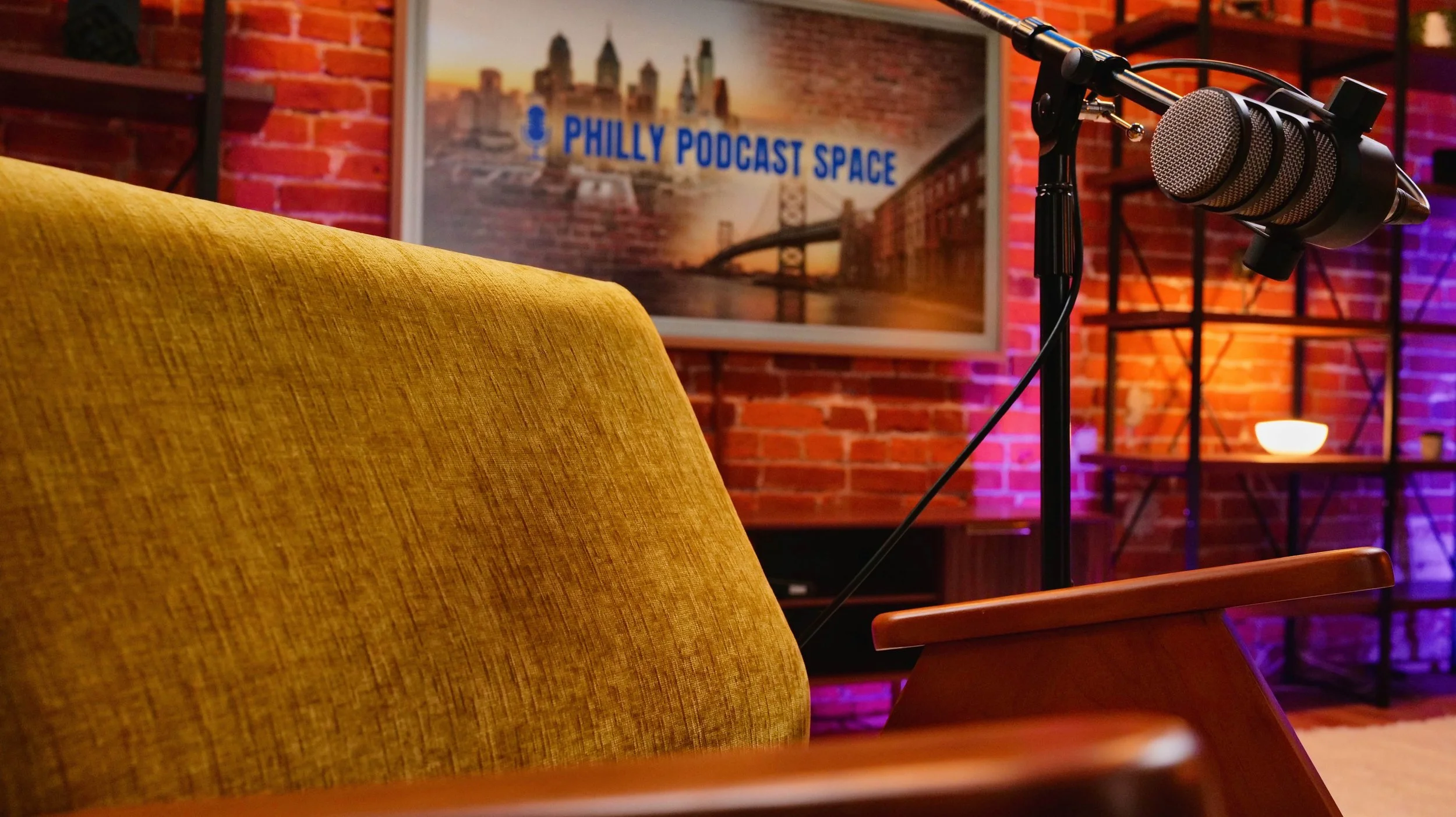 Podcast recording studio with a microphone on a stand, mustard-colored sofa, orange room lighting, and a framed poster that reads 'Philly Podcast Space' with a cityscape