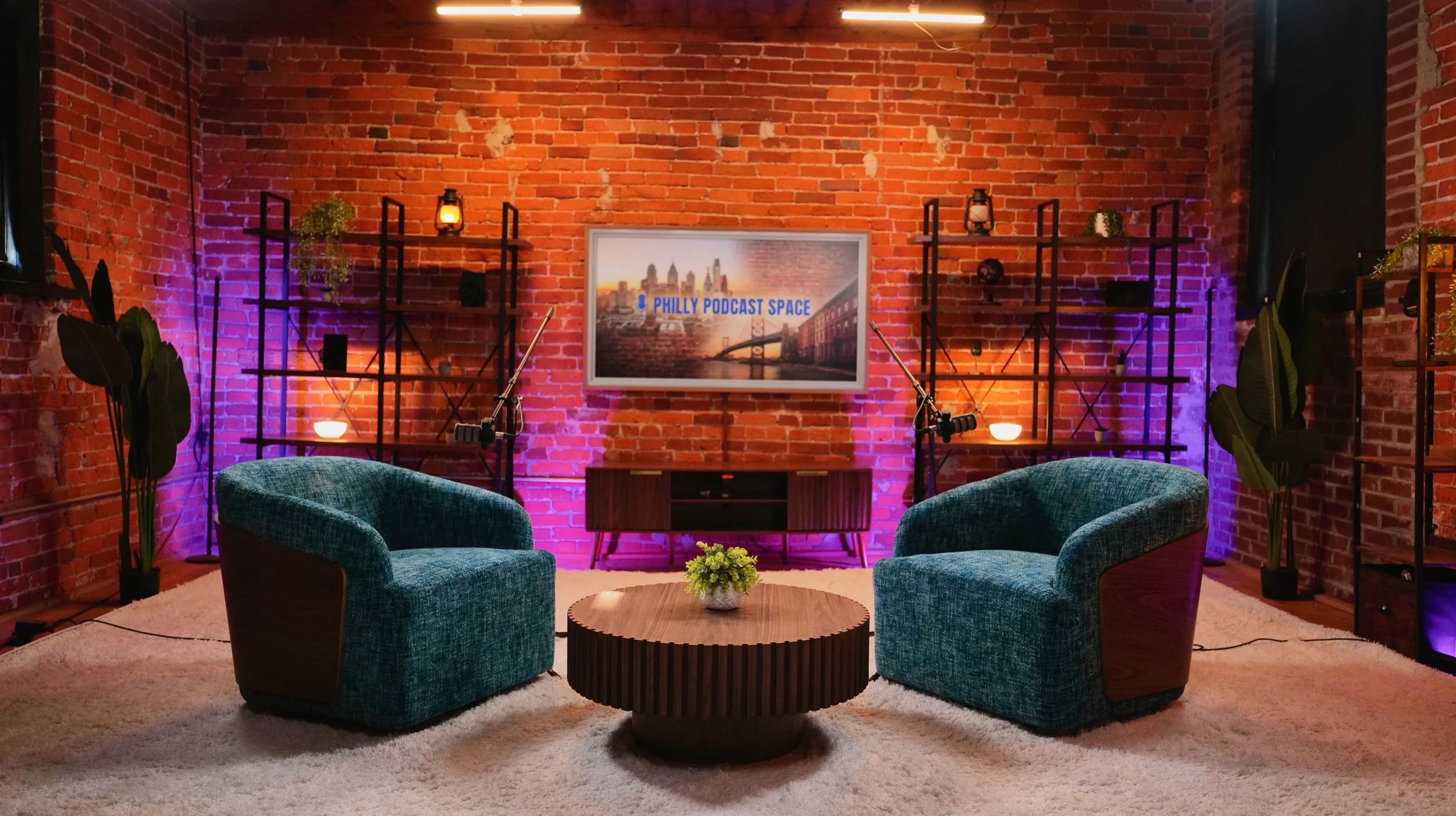 A podcast studio with two green velvet armchairs facing each other, a wooden coffee table with a small flower arrangement, brick walls, shelves with decorative items, potted plants, a TV screen displaying 'Philly Podcast Space', and colorful lighting