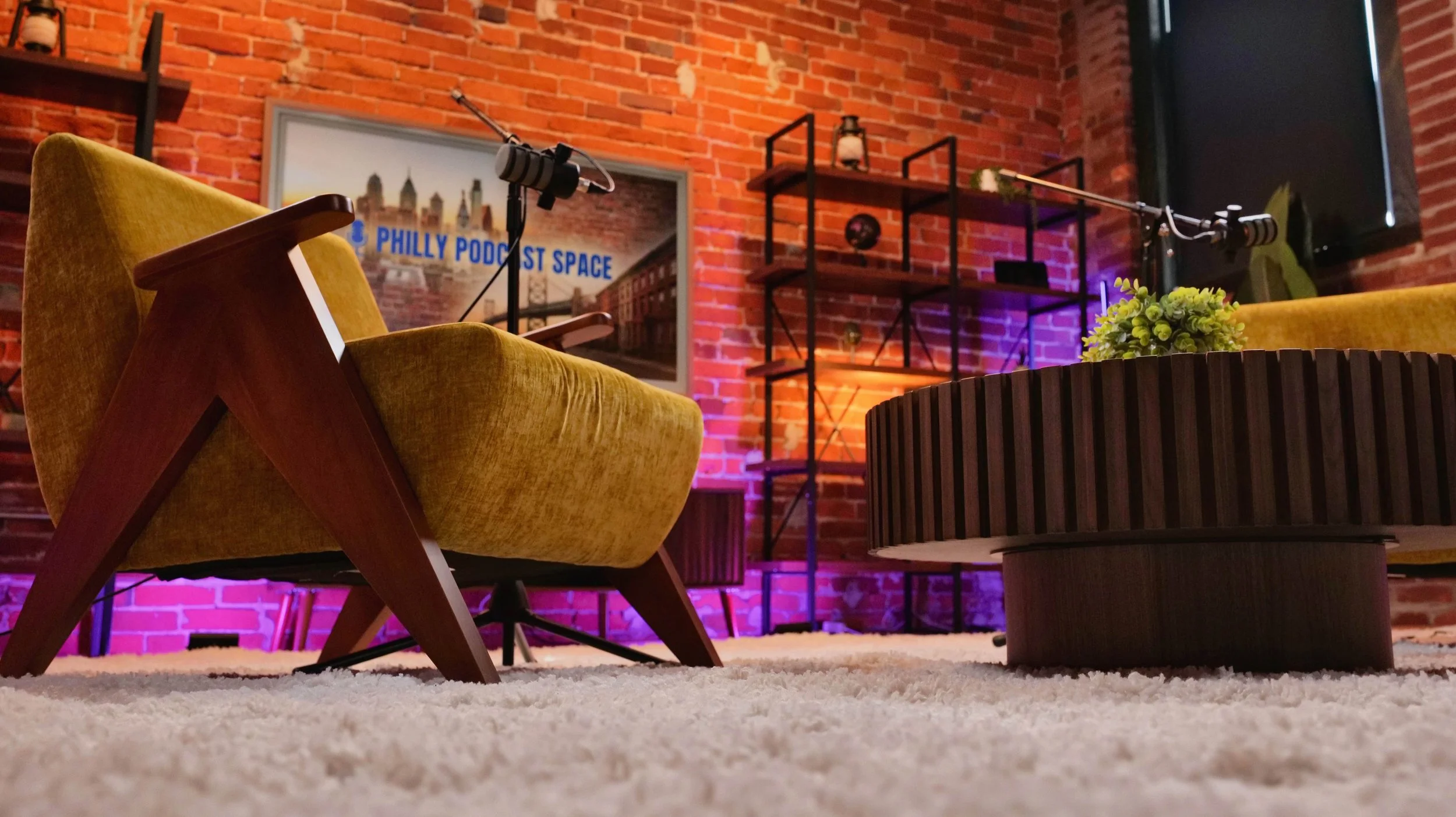 A cozy, modern podcast studio with yellow velvet armchairs, a large round wooden coffee table with a small plant, a brick wall with a framed cityscape picture that reads 'PHILLY PODCAST SPACE,' a black metal shelving unit with decorations, and profes