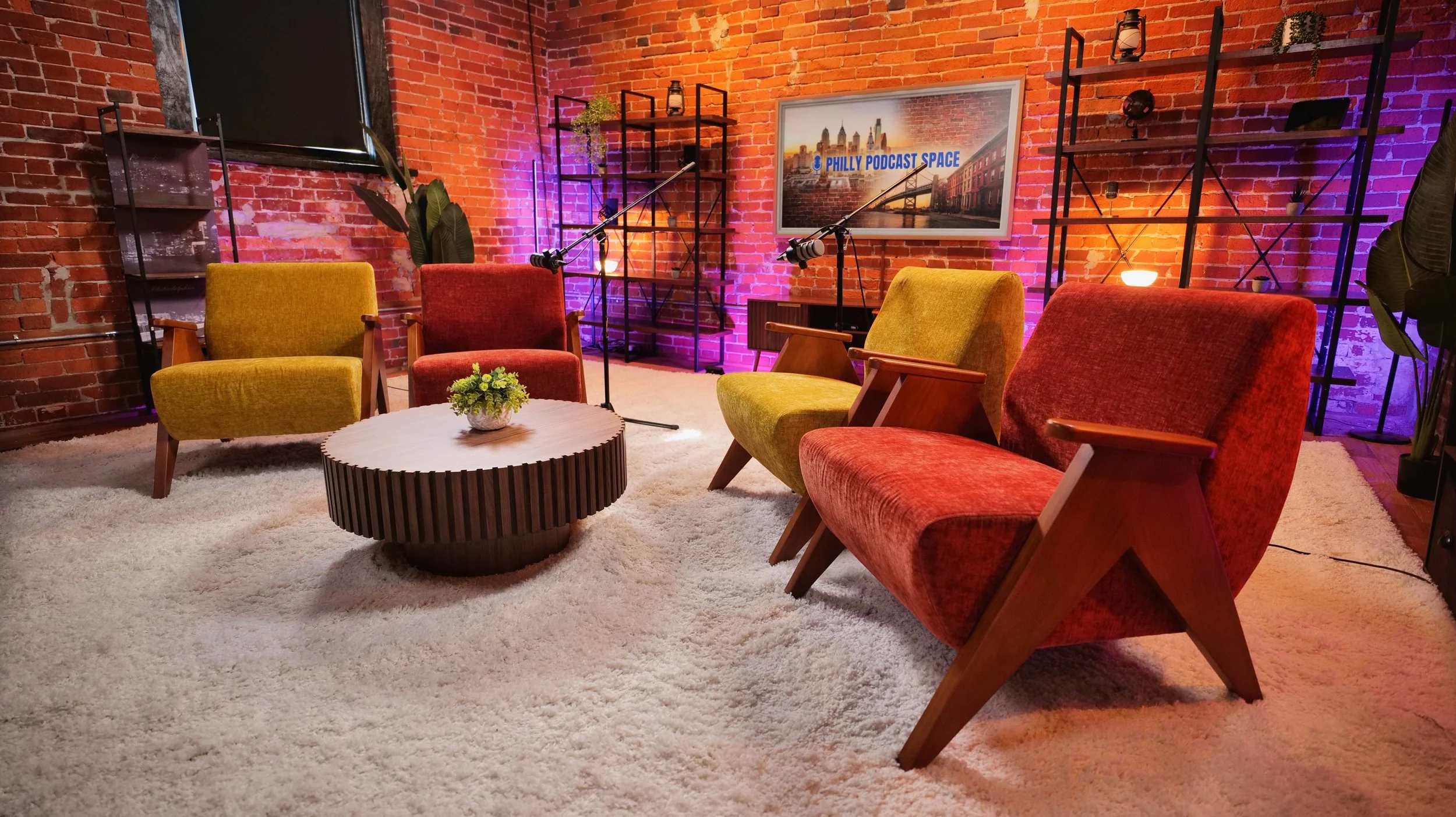 A podcast studio with four colorful vintage chairs, two yellow and two rust-red, arranged around a round wooden coffee table with a small plant, set on a white shag rug. The background features exposed brick walls with shelves, a TV, and a framed pho