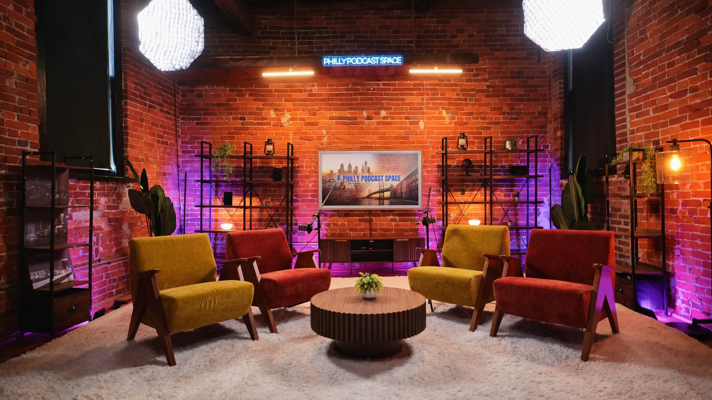 Comfortable seating arrangement with colorful armchairs around a coffee table in a cozy, modern podcast studio with exposed brick walls, decorative shelves, plants, and lighting.