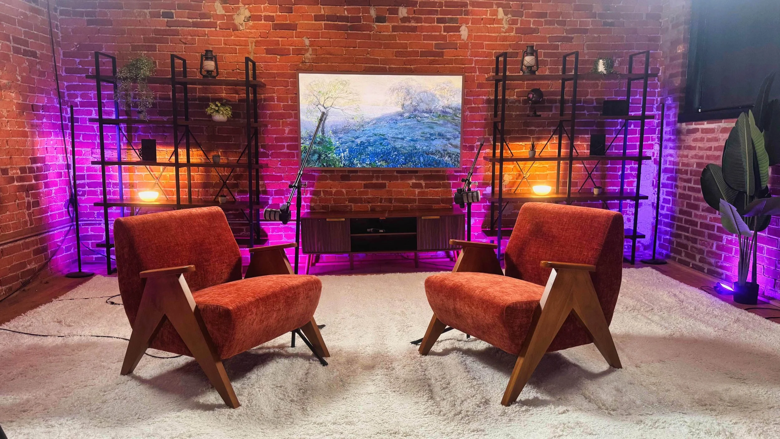 A cozy living room with a brick wall, two orange armchairs, a white rug, and two black shelving units with plants, lit candles, and decorative items, accented with purple and orange LED lighting.