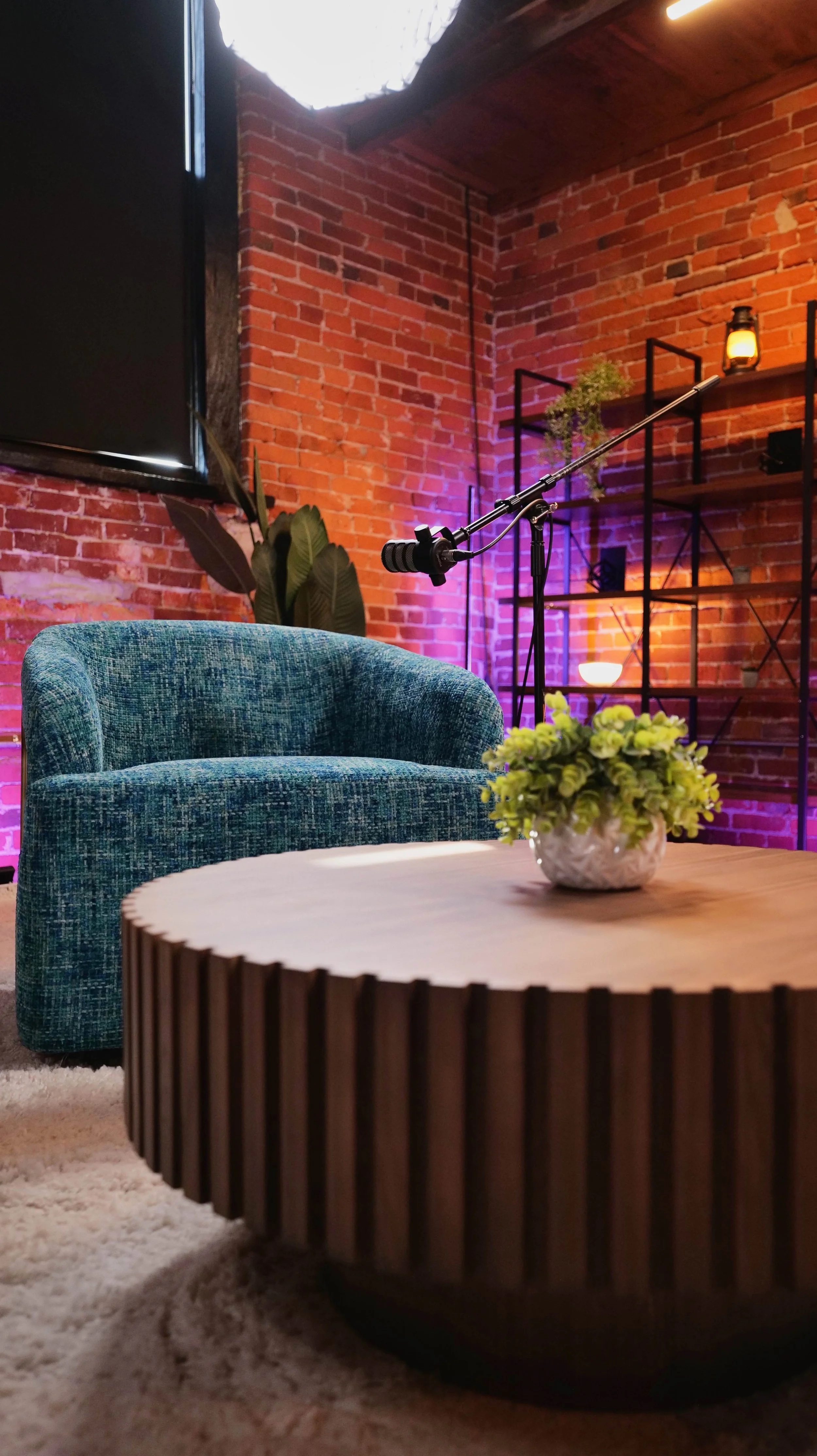 A cozy modern living room with a green upholstered armchair, a round wooden coffee table with a potted green plant, a brick accent wall, and a black metal shelving unit with decorative lighting in the background.