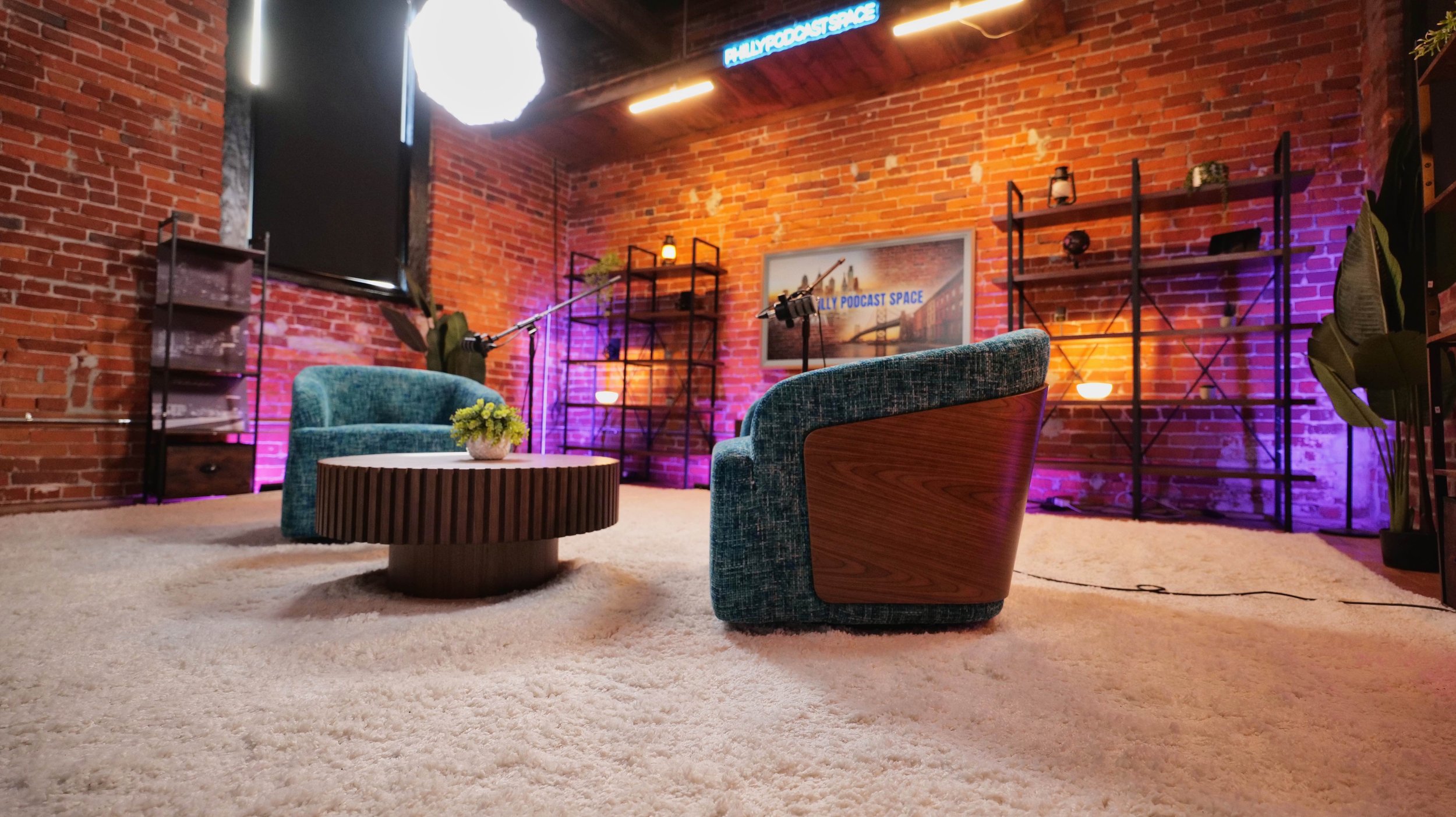 Interior of a podcast studio with brick walls, two blue velvet chairs, a round wooden table with a small plant, and metal shelving units lit with purple and orange lighting. A framed picture reads 'Billy Podcast Space', and a large softbox light is v