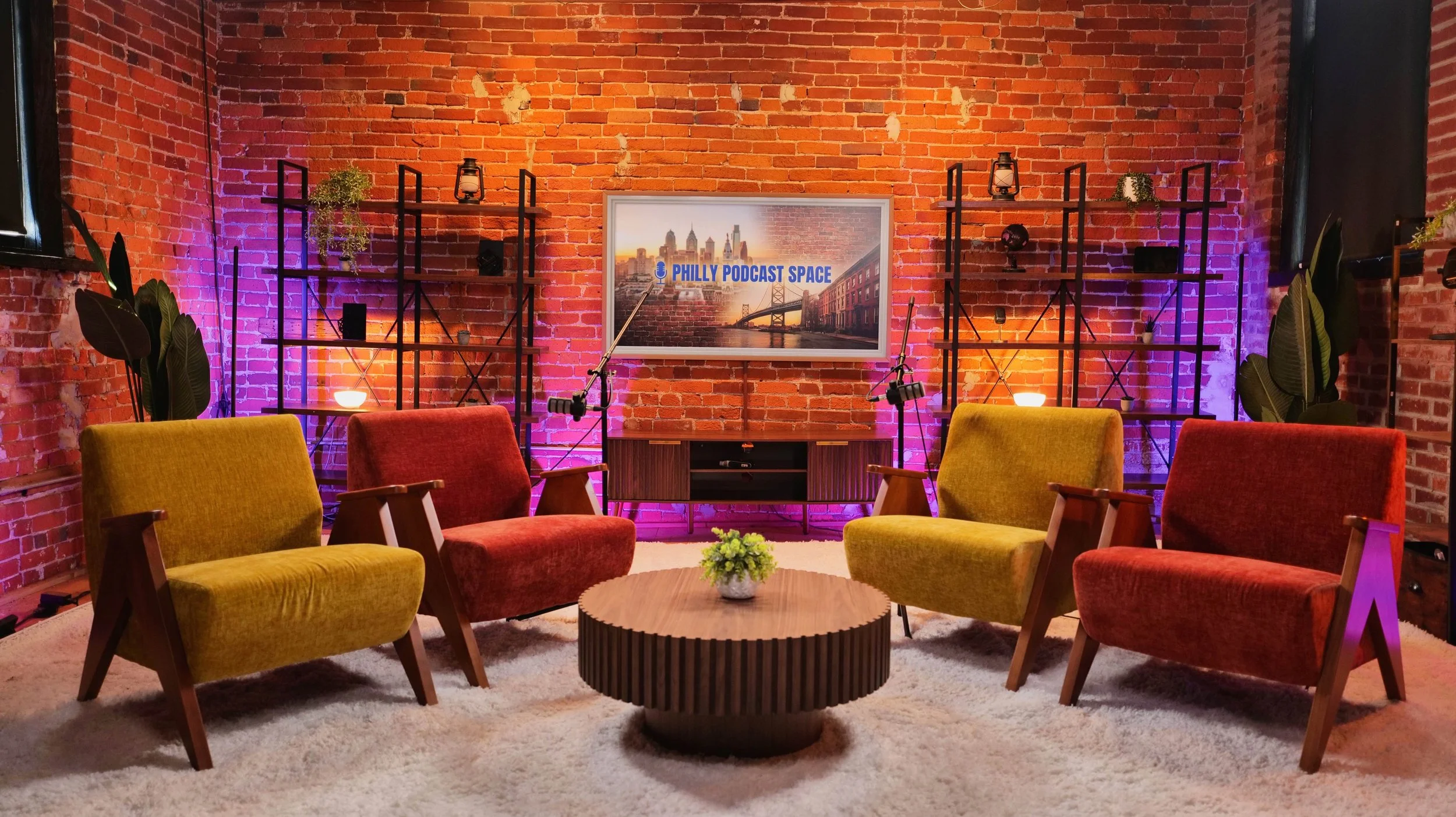 A cozy studio with a brick wall backdrop, featuring four mid-century modern armchairs (two yellow and two red) arranged around a round wooden coffee table with a small green plant. Behind the chairs, there are two black shelving units with decorative