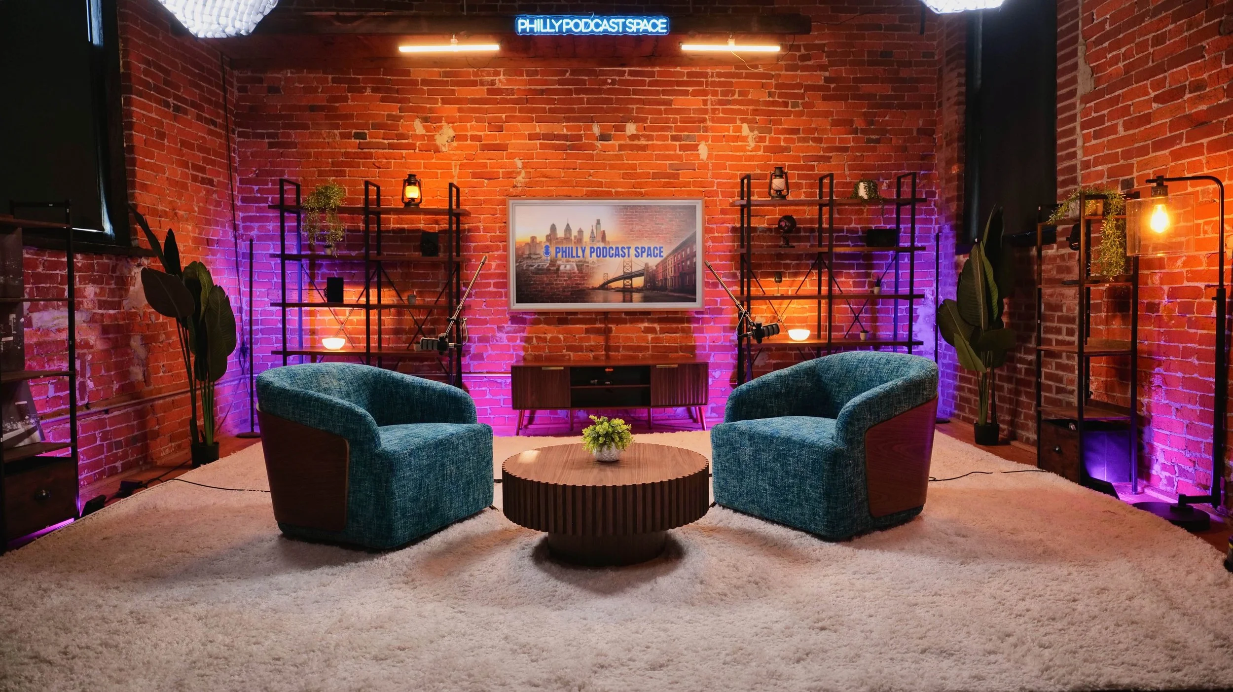 A podcast studio with two teal chairs, a round wooden table with a small plant, brick walls, shelves with decor, purple and orange lighting, and a TV screen displaying 'PHILLY PODCAST SPACE' with a city skyline background.