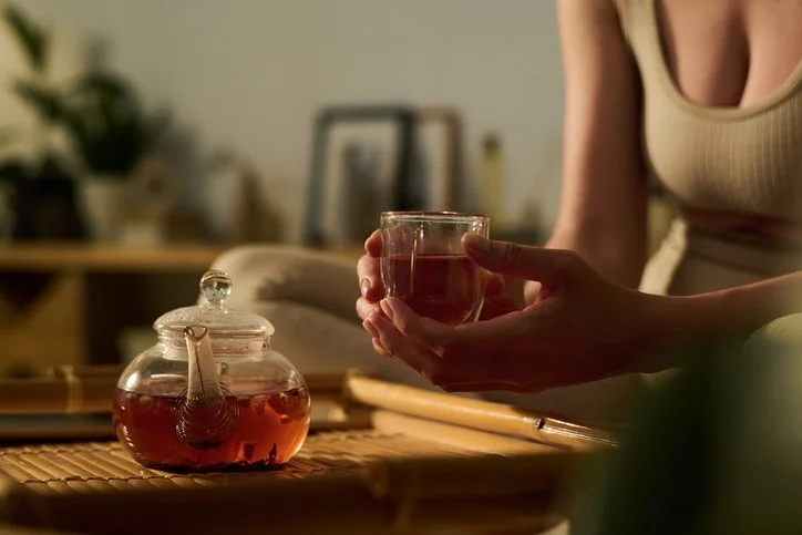 Woman holding a glass cup of herbal tea beside a teapot in warm evening light, representing a calming nighttime ritual for nervous system support.