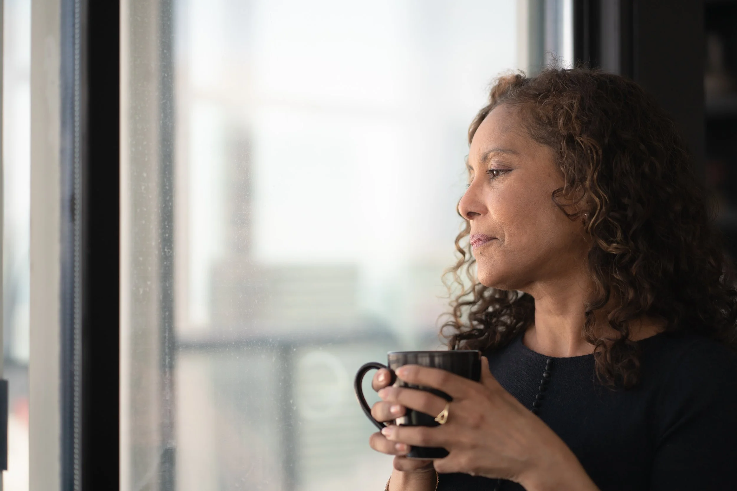 Woman holding a mug and looking thoughtfully out a window.