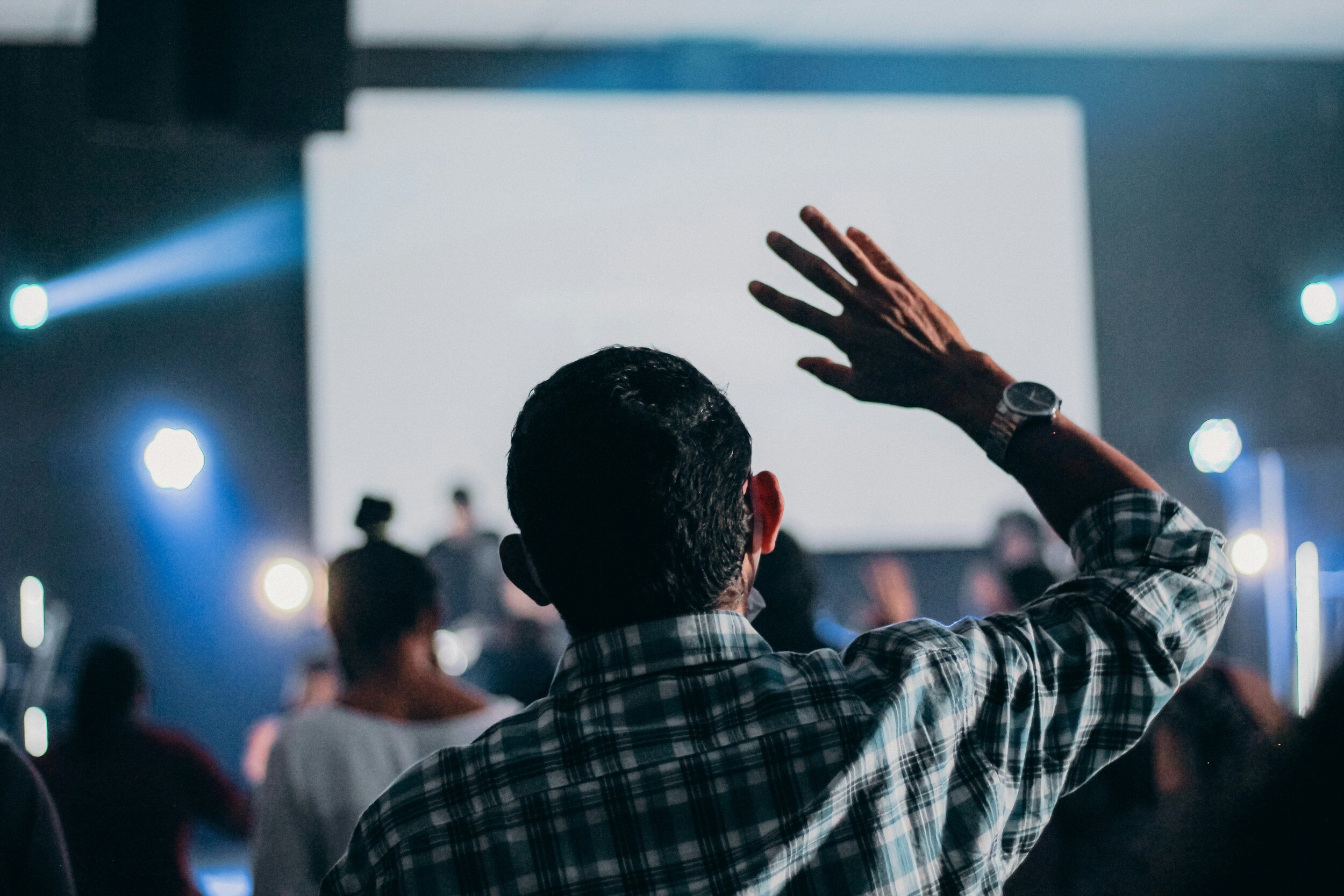 A person with short dark hair wearing a plaid shirt raising his right hand towards a large blank screen in a dimly lit room filled with other people. The scene appears to be a presentation or seminar with stage lighting.