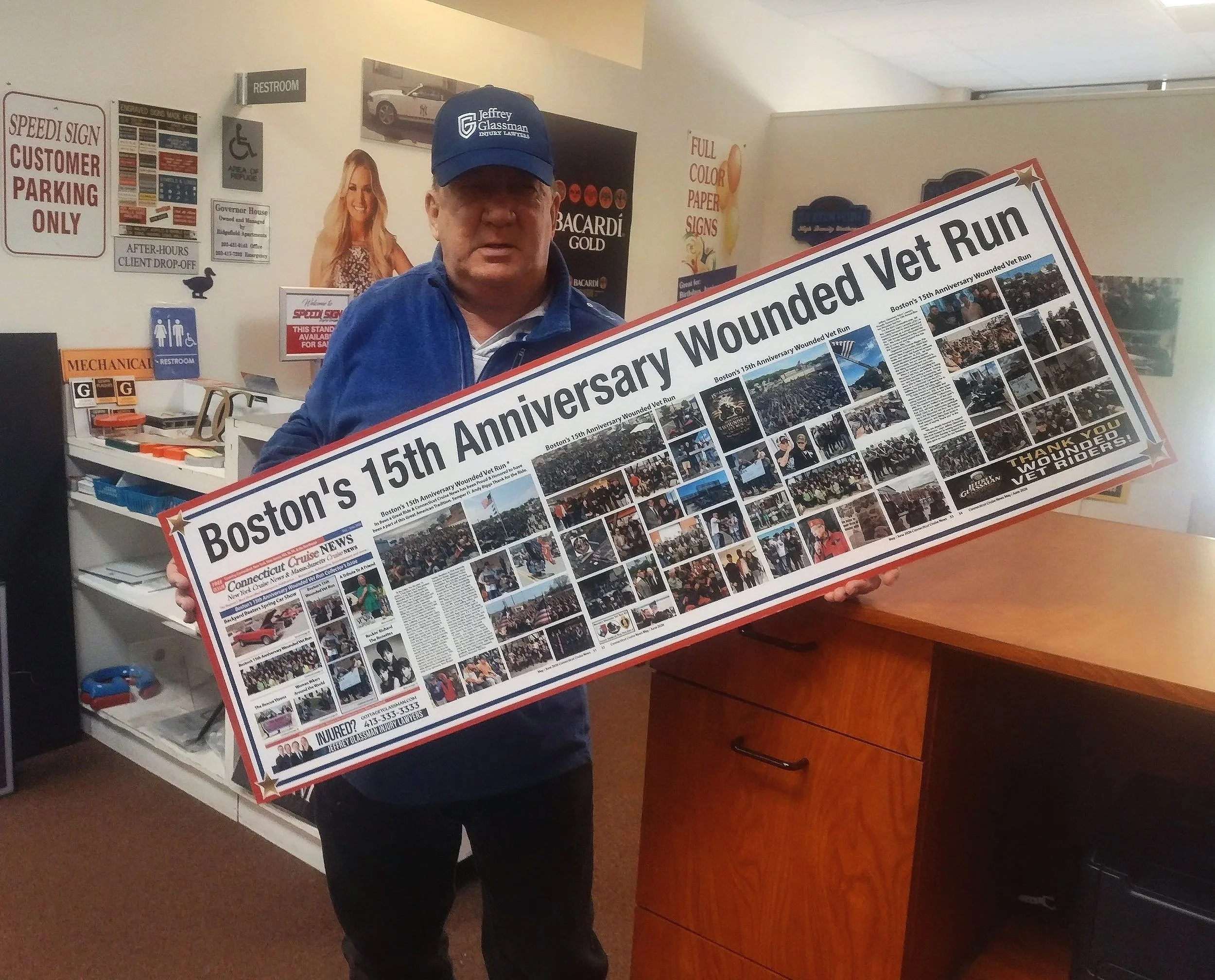 A man in a blue cap holding a sign for Boston's 15th Anniversary Wounded Vet Run on which is featured lots and lots of photos of the run
