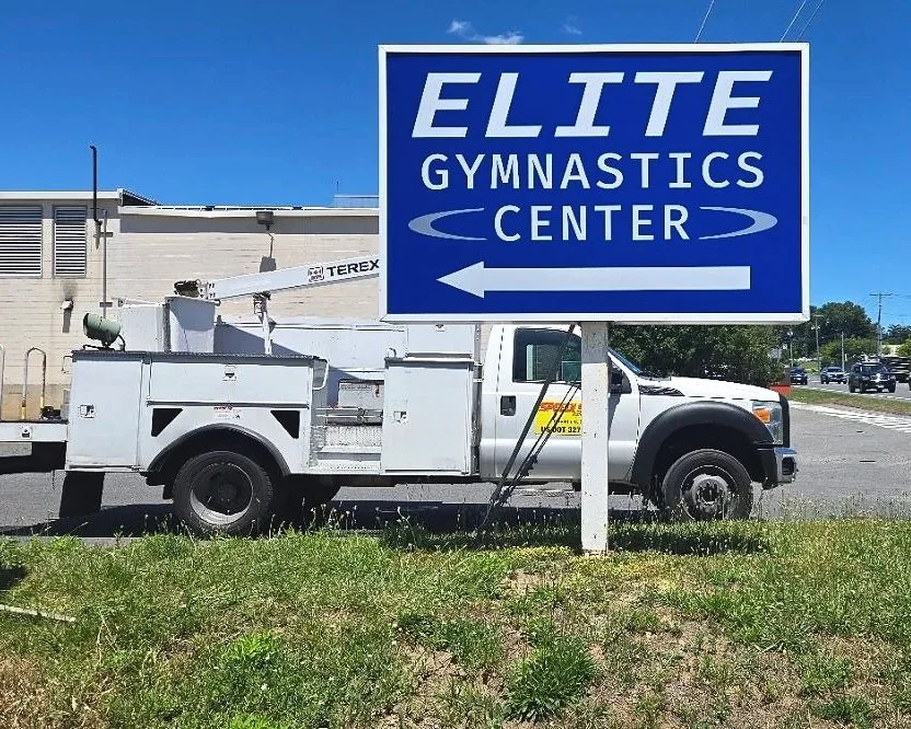 A sign for Elite Gymnastics Center - white text on a blue background. Sign is on a grass verge and parked behind it is a Speedi Sign truck.