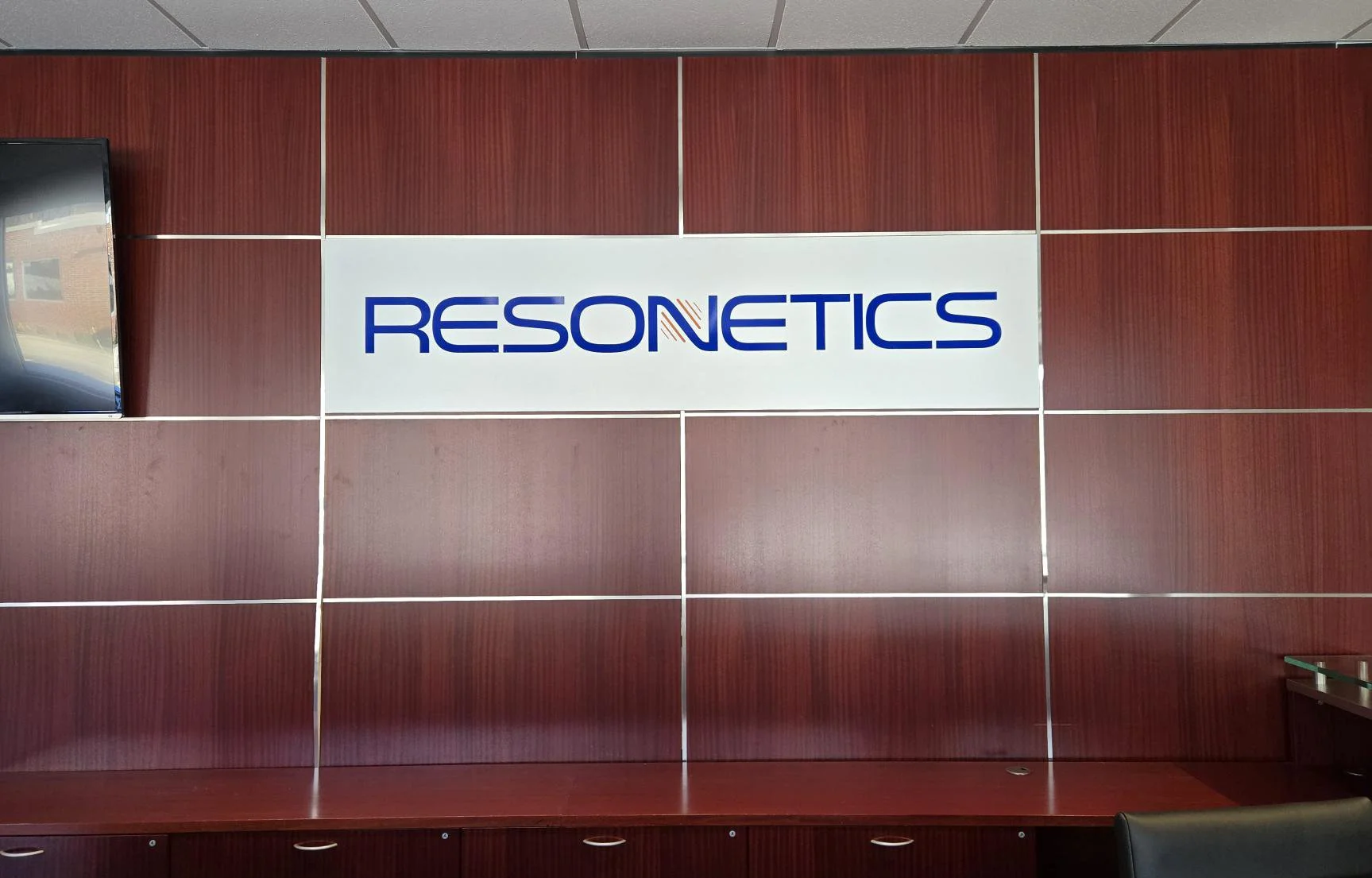 interior sign at Resonetics office - blue modern font on white background on a wall with mahogany colored wood paneling 