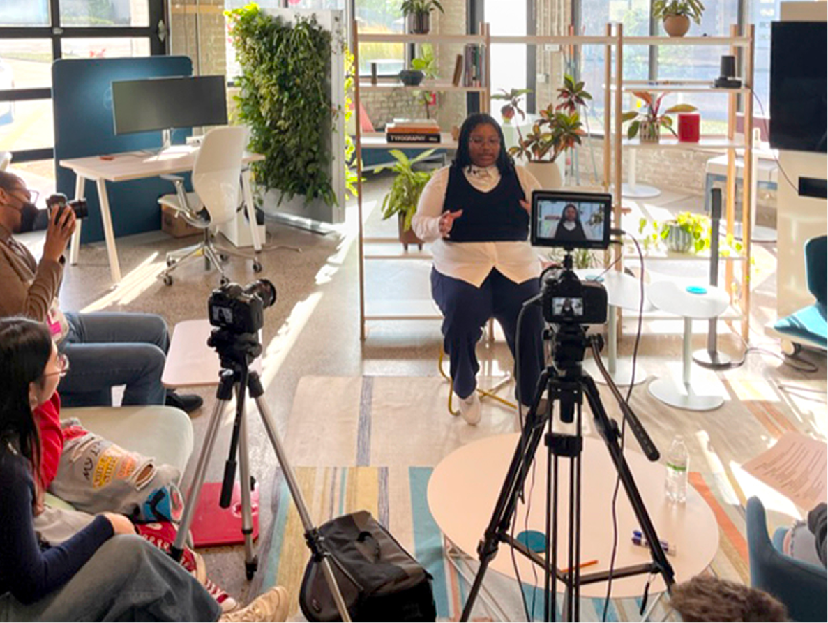 A woman speaks in front of a camera with an audience in a bright, plant-filled office or studio setting, with large windows and various furniture and plants in the background.
