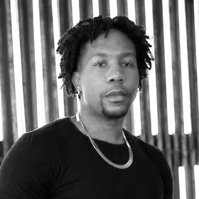A young man with curly hair and earrings, wearing a black shirt and a silver chain, standing in front of vertical wooden slats.