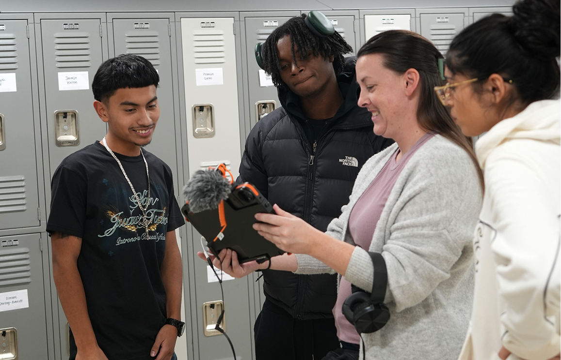 A group of four diverse students, two women and two men, smiling and looking at a recording device held by one of the women in a school hallway with gray lockers.