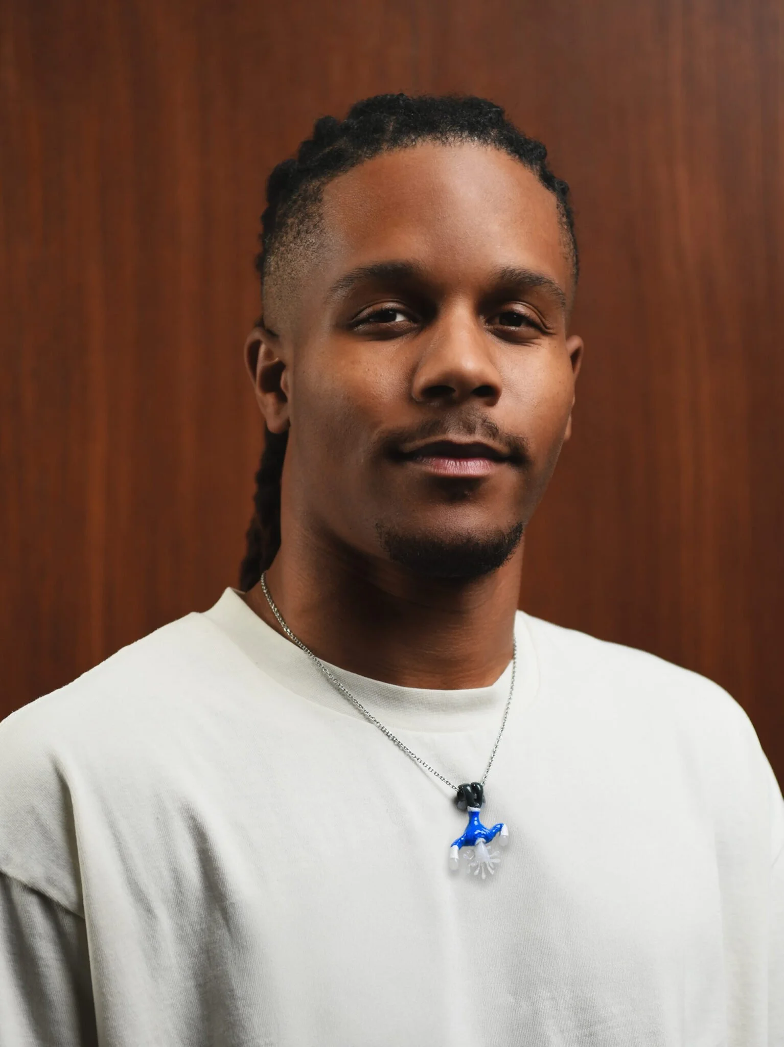 Close-up of a young man with dark skin, black hair styled in dreadlocks, wearing a white shirt and a necklace with a blue and white ornament.